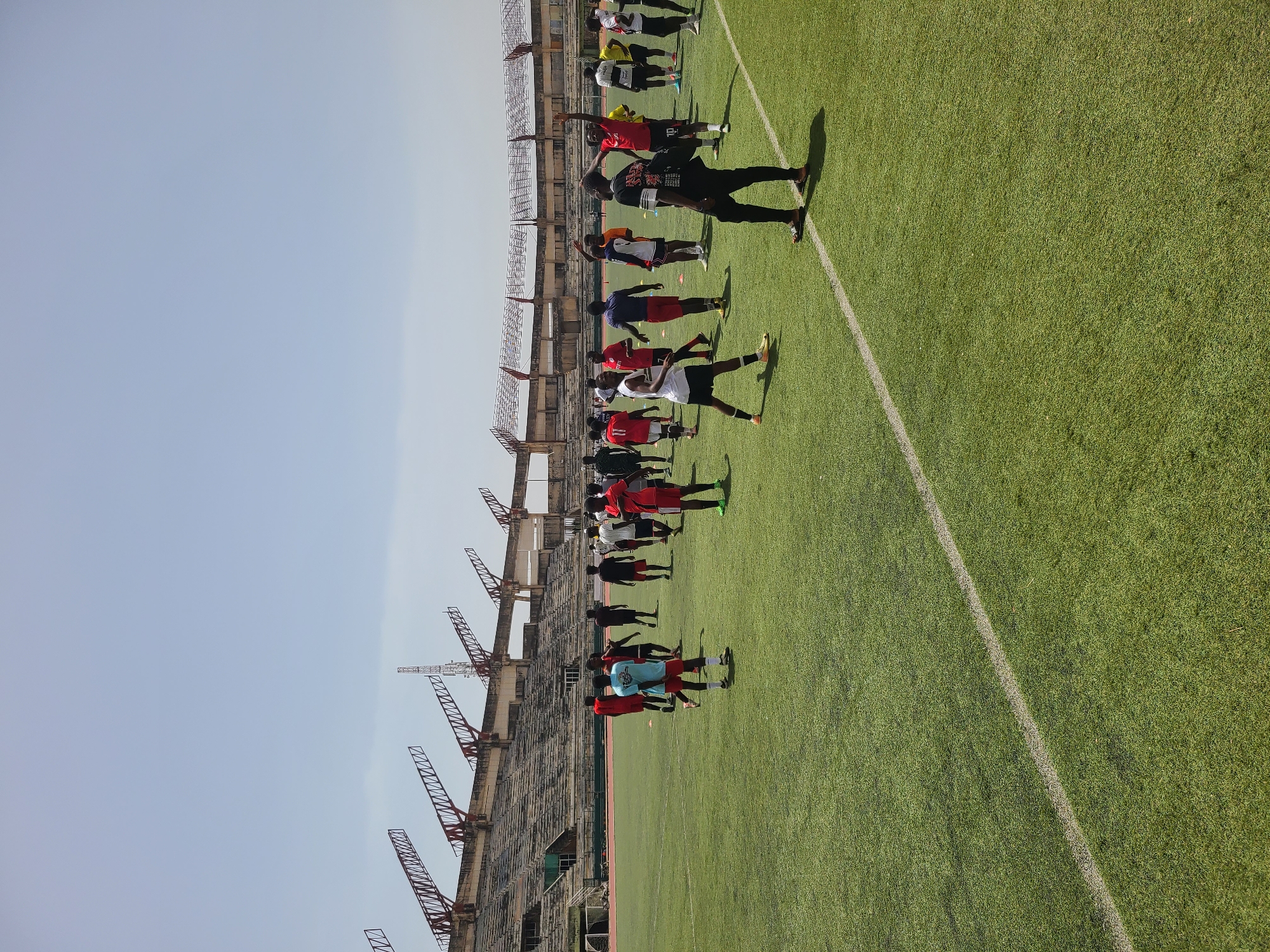 The image depicts a group of people lined up on a green field next to a stadium under a clear sky. The stadium is a concrete structure with a steel frame. The people are wearing a variety of clothing, including red, white, and black shirts. Some of them appear to be stretching or warming up. The field has a white line running along the edge of the frame. The location is New Stadium, Nigeria.