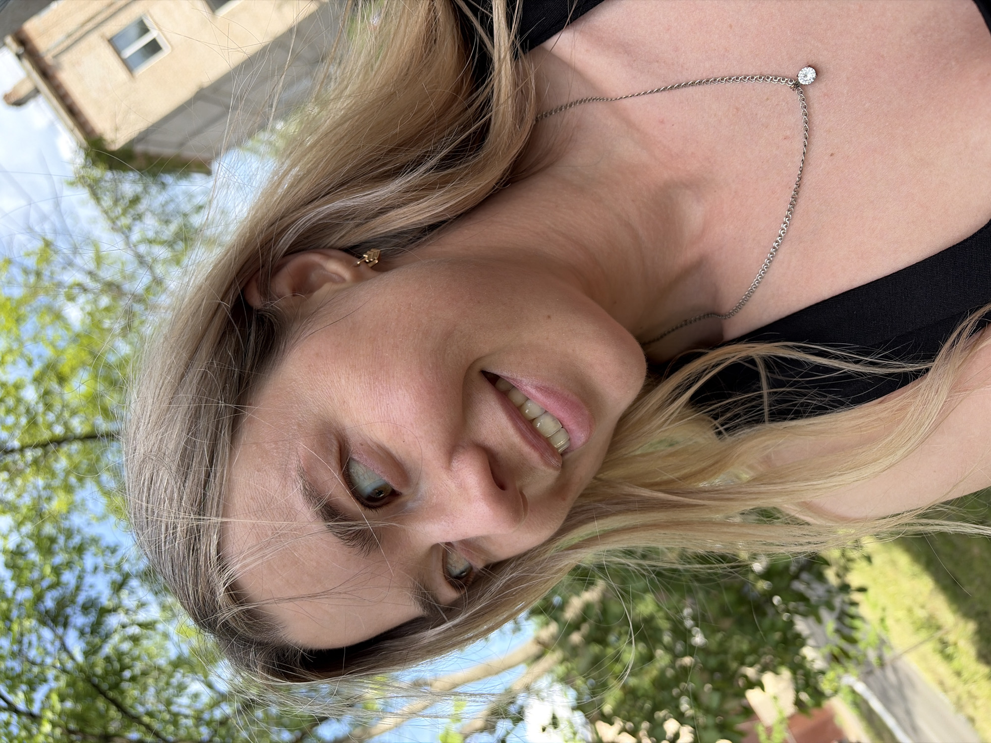 A young woman is photographed from a low angle, seemingly outdoors. She is fair-skinned with blonde hair and wears a black top and a silver chain necklace with a diamond-like pendant. Part of a building with a window is visible in the background, along with trees and a blue sky. The location is not explicitly identifiable as Chaikovsky, Russia based solely on the visual information present in the image.