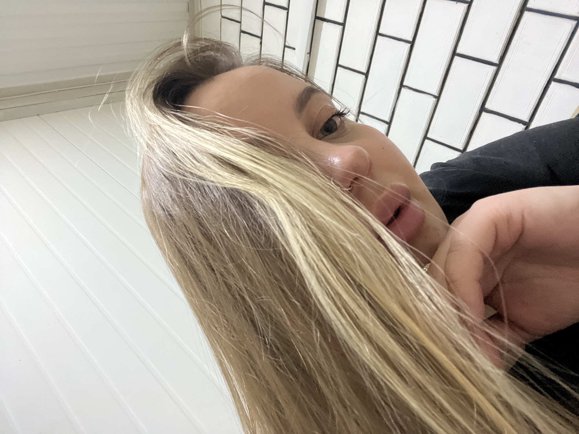A close-up, angled shot of a blonde woman indoors. The background features two types of wall paneling: horizontal white panels on the left and vertical white panels with black trim on the right. The woman's face is partially obscured by her long blonde hair. A hand is visible near her face. The image does not provide enough information to confirm the location or any activity in Novopokrovskaya, Russia.