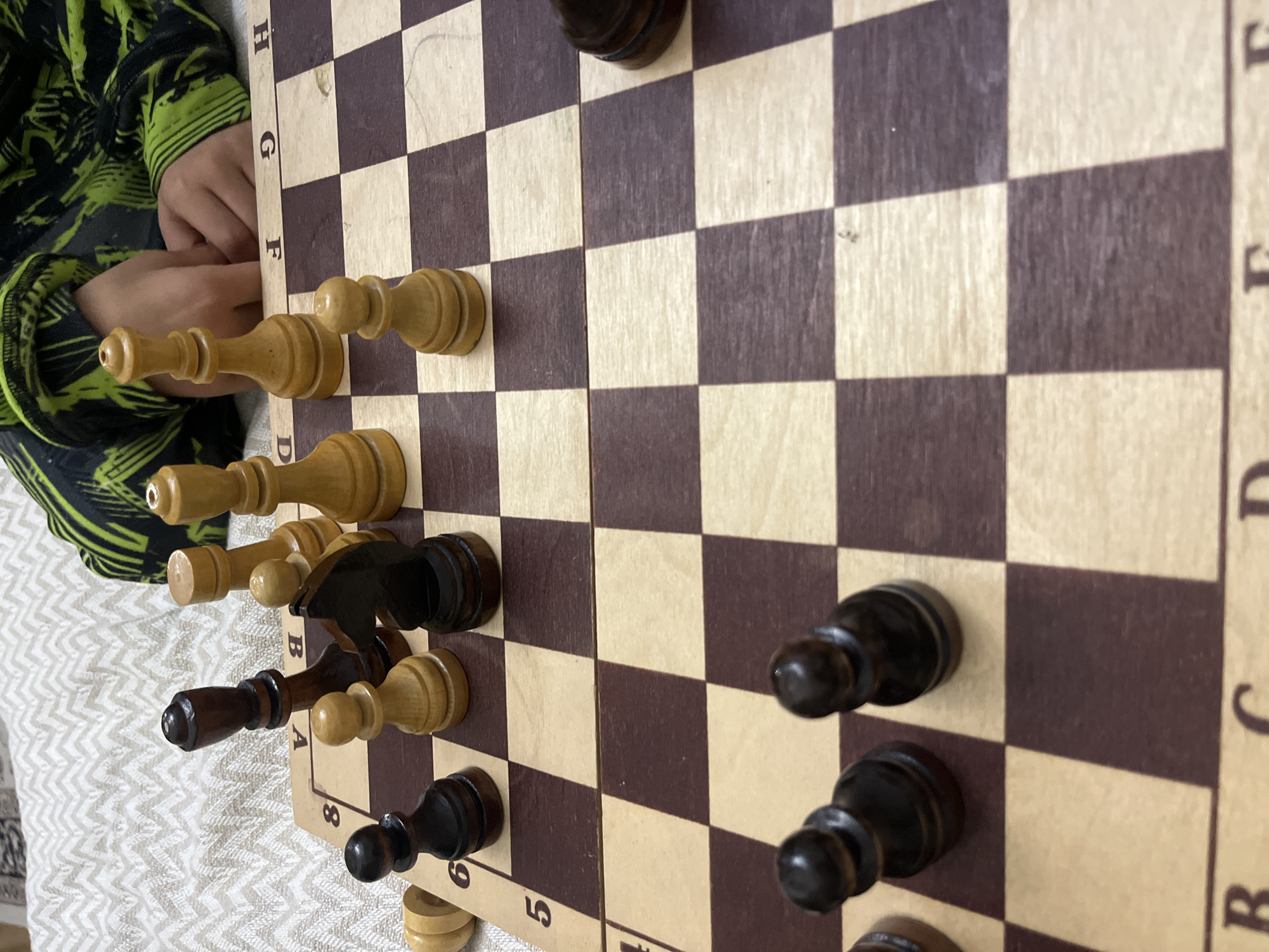 A close-up shot depicts a chessboard with chess pieces in mid-game. The pieces are made of light and dark wood. White pieces occupy the top portion of the image, with a black knight prominent in the center. Several black pawns are positioned at the bottom. A child's hand is visible in the upper left corner, resting on the board's edge. The coordinates A-H and 1-8 are visible on the board's periphery. No information is provided to infer anything about the city Kaspiysk, Russia.