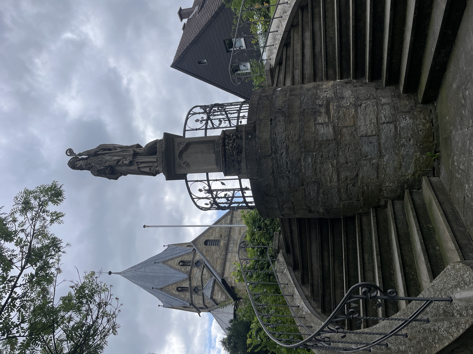 The image shows a stone statue of a man in a robe and headdress holding a staff, standing on a pedestal. The pedestal has writing on it. Two curved stone staircases with wrought iron railings lead up to the pedestal from the foreground. Behind the statue and stairs is a stone church with a tall, pointed grey roof and a spire. To the right of the church is a dark-colored house with a pitched roof and a chimney. The scene is set against a cloudy sky. The location is the St. Liborius monument in Salzkotten, Germany.
