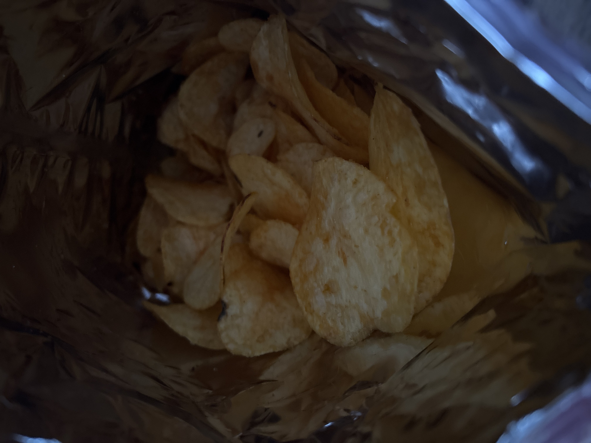 The image depicts a close-up view inside a partially empty bag of potato chips. The chips appear to be of a standard variety, with a light golden color and some visible seasoning. The bag is made of a reflective, metallic material. The lighting is dim, with some areas appearing darker due to shadows. There is no indication of the image's location.