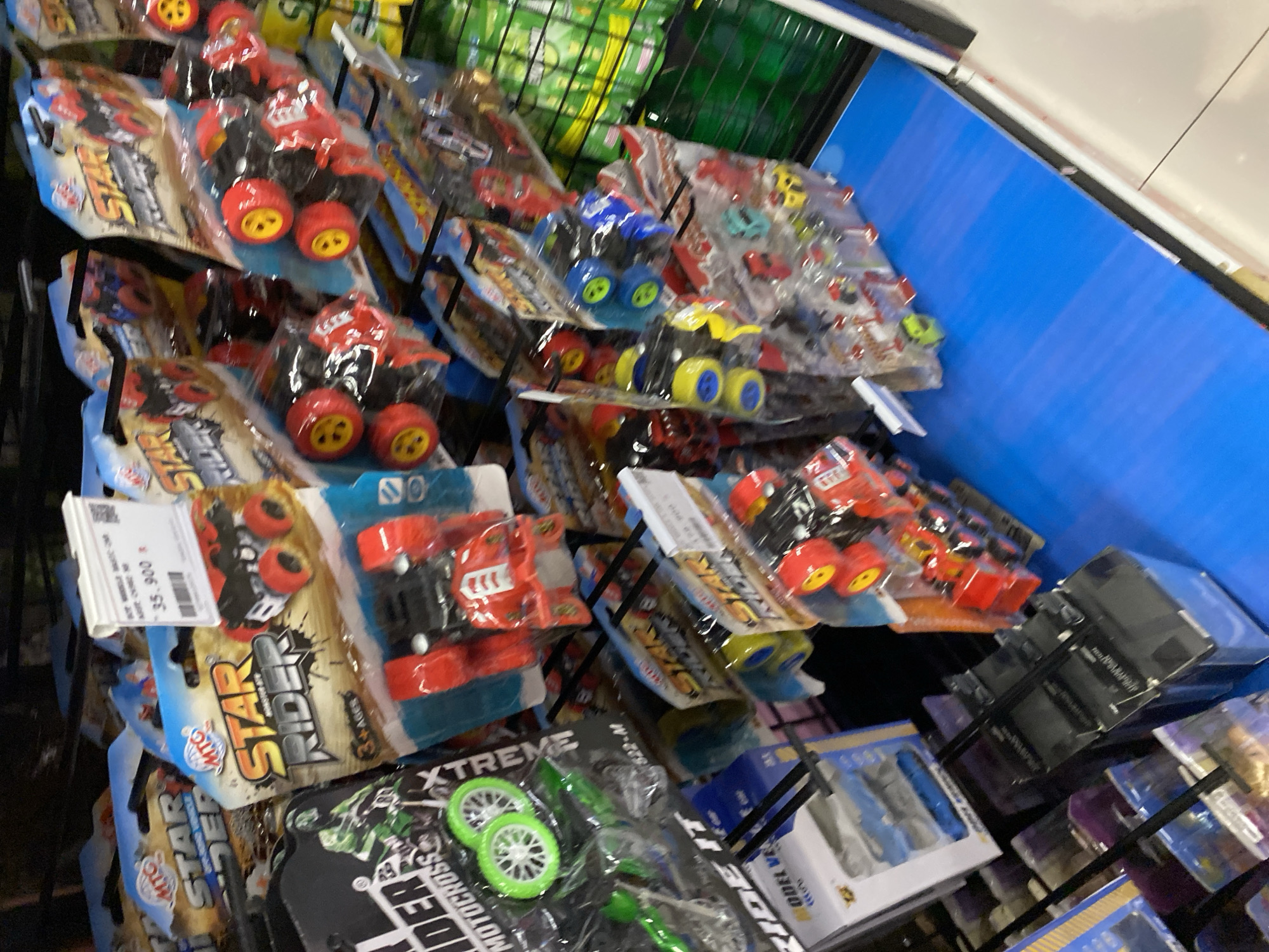 The image shows a shelf display of children's toys in a store, likely a supermarket or toy shop. The toys are packaged in plastic blisters and cardboard, predominantly featuring toy cars and motorcycles. Some prominent brands visible include "STAR RIDER" and "MTG". The toys are in various colors, with red, blue, and green being noticeable. A price tag with the amount "35.900" is visible on one of the packages. The background suggests a retail environment with shelving and other products. There are no people visible in the image. The image is an indoor shot with artificial lighting.