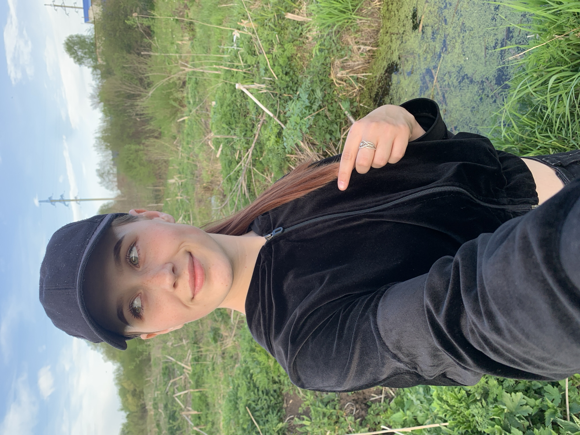 A young woman with fair skin, freckles, and auburn hair is outdoors, wearing a black baseball cap and a black velvet jacket. She is smiling slightly and looking towards the camera. Her hair is pulled back, revealing her face. On her finger is a silver ring. The background includes green grass, shrubs, a body of water covered with algae, and power lines in the distance. The location cannot be determined as Фоки, Russia based on the information in the image.