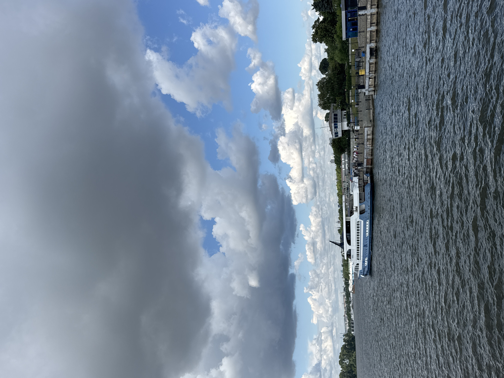 A riverboat is docked at a pier on a large river in Kazan, Russia. The sky is partly cloudy with dark storm clouds in the upper left and lighter clouds and blue sky in the upper right. The river is choppy with small waves. The pier is made of concrete and has a railing along the edge. Several people are standing on the pier, some near the boat and others further down. There is a building with a tower-like structure on the pier, and beyond that, a line of trees and buildings along the riverbank. A bridge can be seen in the distance on the left side of the image. The boat is white and blue with several windows and appears to be a passenger vessel.