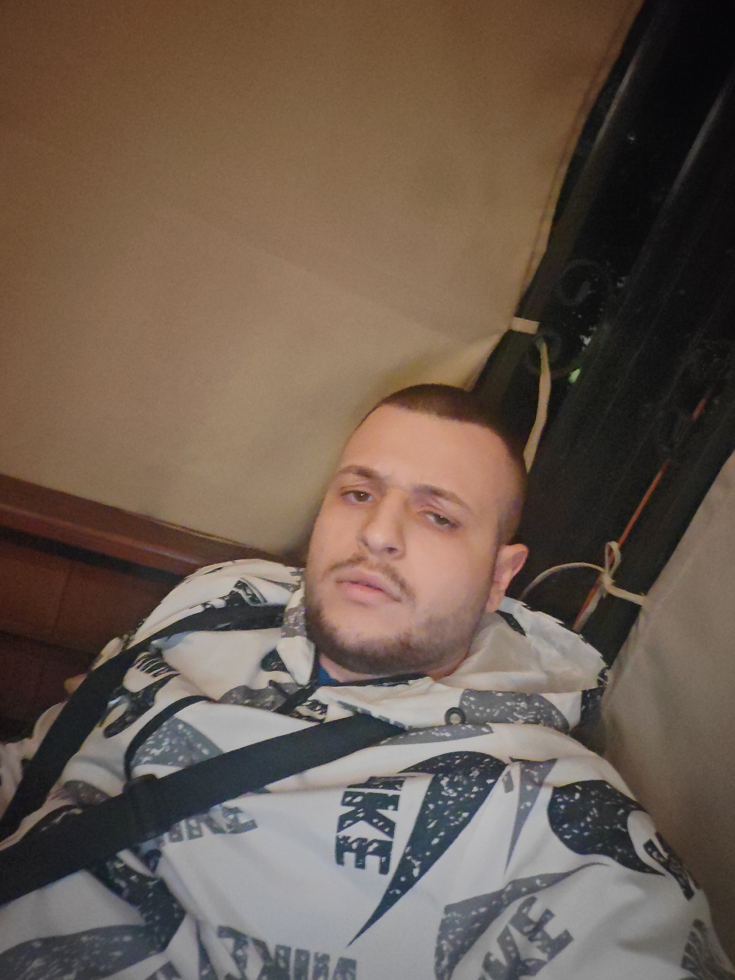 A young man with short, dark hair and a beard is lying down, facing the camera. He is wearing a white hooded jacket with a black and grey abstract pattern. A black strap, possibly from a bag, is visible across his chest. The background consists of brown upholstery and dark, vertical panels with some rope ties. The image appears to be a selfie taken indoors. The man is not in a recognizable location within the city of Burgas, Bulgaria.