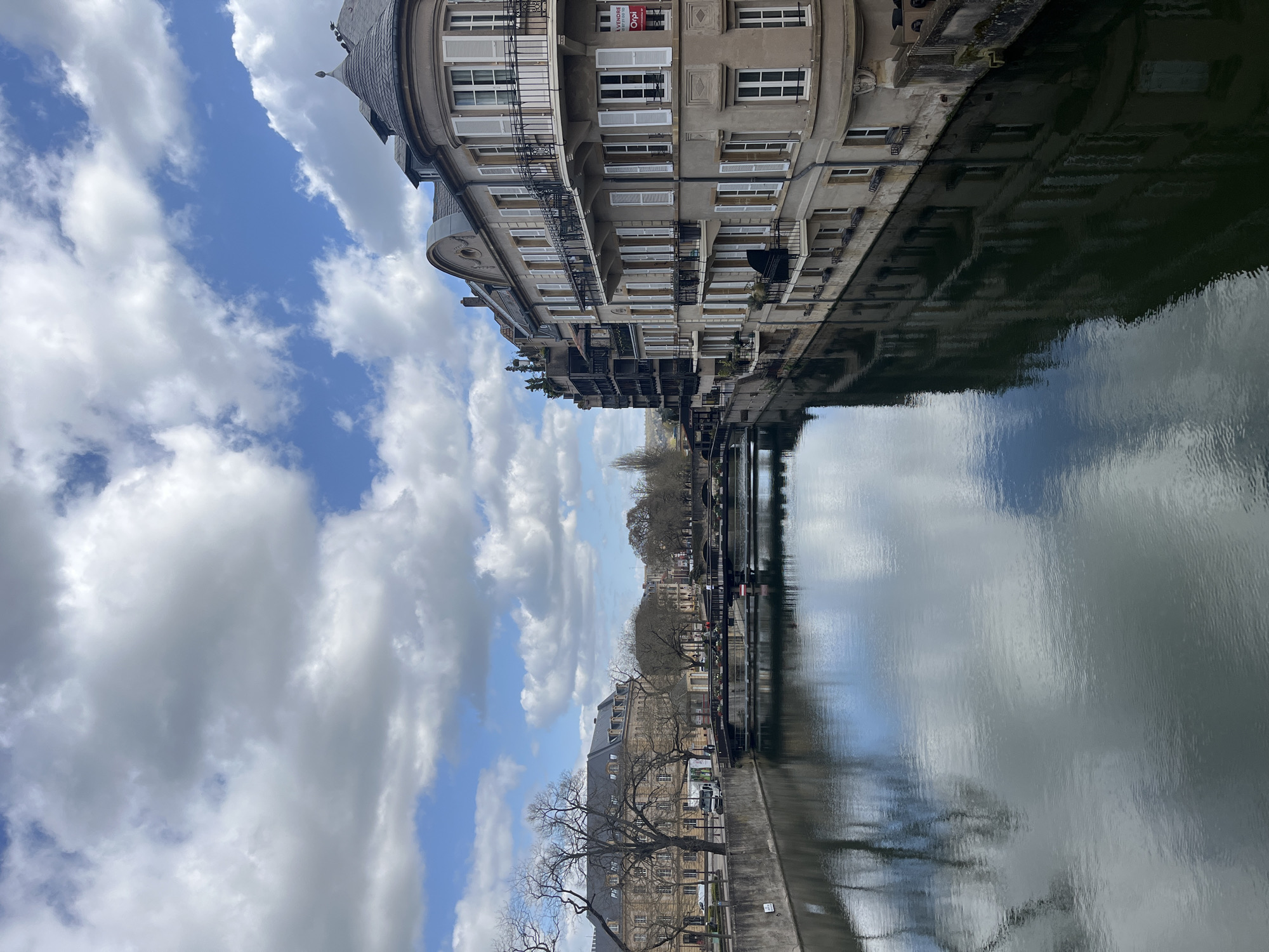 The image shows a canal scene in Metz, France, with buildings lining the waterway reflected in the water. The buildings have multiple floors, light-colored shutters, and balconies. A "A vendre" sign is visible on one of the buildings. A bridge crosses the canal in the distance. The sky is blue with numerous white clouds. Bare trees line the opposite bank of the canal. The water is reflecting the sky and buildings.