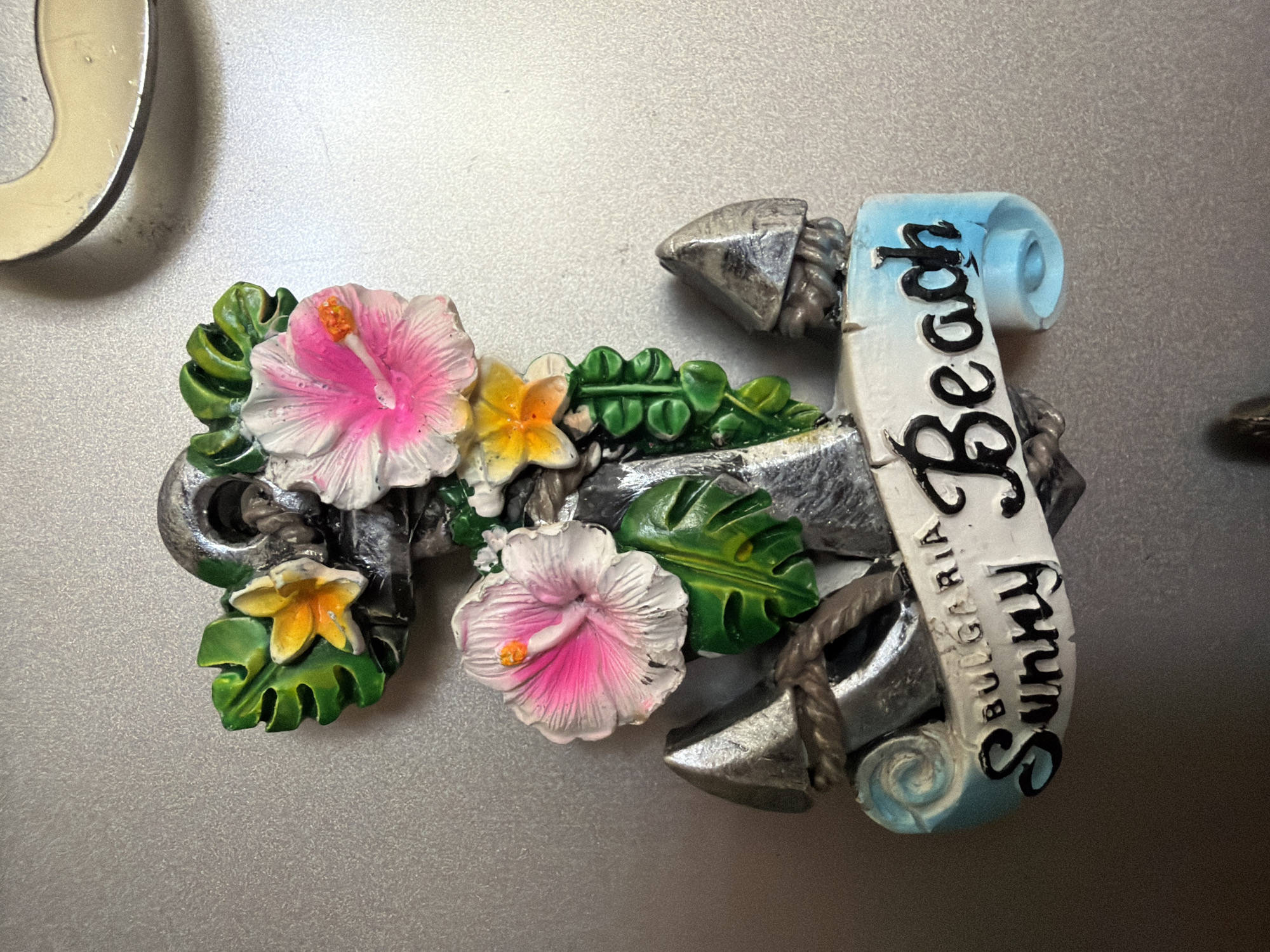 The image shows a close-up of a decorative magnet shaped like an anchor adorned with flowers and leaves. The anchor is metallic gray, with a rope detail wrapped around it. Two pink and white hibiscus flowers and two yellow flowers are attached to the top of the anchor, along with green tropical leaves. Below the anchor, a white banner with blue accents unfurls, displaying the words "BULGARIA" and "Sunny Beach" in black lettering. The magnet is placed on a textured, light gray surface, possibly a refrigerator or a similar metallic object. The lighting is even, highlighting the details of the magnet. There is no indication of any specific location in the city of Pazardzhik, Bulgaria, beyond what is written on the magnet itself.