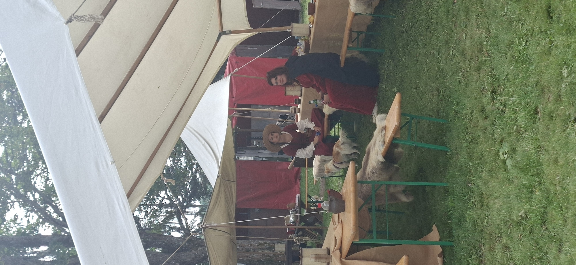 The image depicts an outdoor scene under a large white tent, possibly at a historical reenactment event, set in the city of Turku, Finland. Two young women are seated on a wooden bench. The woman on the left wears a straw hat and a reddish-brown tunic over a white blouse, her hair pulled back. The woman on the right wears a dark cloak over a long red dress, with her hair down. Both are looking towards the camera. In front of them, on a wooden table, are several objects, including a dark jug and a bottle of water. Two small dogs are present; one is standing near the bench on the left, and the other is partially visible under a table with a fur-covered bench in the foreground. The ground is covered in green grass. In the background, there are additional tents with red fabric drapes. The overall atmosphere suggests a rustic, historical setting.