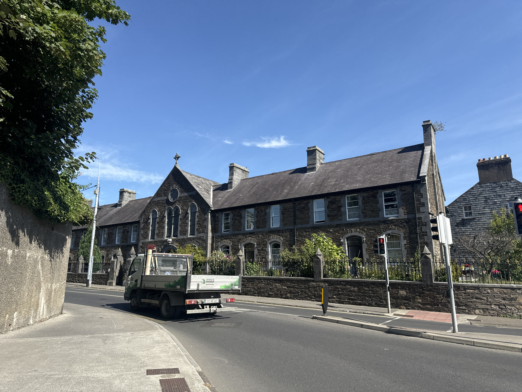 The image captures a street view in Waterford, Ireland, on a bright, sunny day with a clear blue sky and scattered wispy clouds. On the left, a textured grey stone wall partially obstructs the view of a lush green tree with dense foliage. A paved sidewalk runs along the wall, curving slightly to the right.

Dominating the scene is a long, dark stone building with a slate roof, featuring multiple chimneys. The building exhibits arched windows and a central section with a pitched roof topped by a cross. A black metal fence with decorative posts runs along the front of the building, behind which are various green shrubs and some flowering plants.

In the foreground, a light-colored flatbed truck, with its tailgate down, is parked on the right side of the road, facing away from the viewer. The truck appears to be carrying gardening or construction materials, including what looks like white planks and some green sheeting. The truck's license plate reads "222-KK-407."

A traffic light, showing a red pedestrian signal, is visible to the right of the truck. The road is asphalt, with white lane markings. Another building, with brickwork and a grey roof, is partially visible to the right of the main stone building. The overall impression is one of a well-maintained urban environment bathed in sunlight.