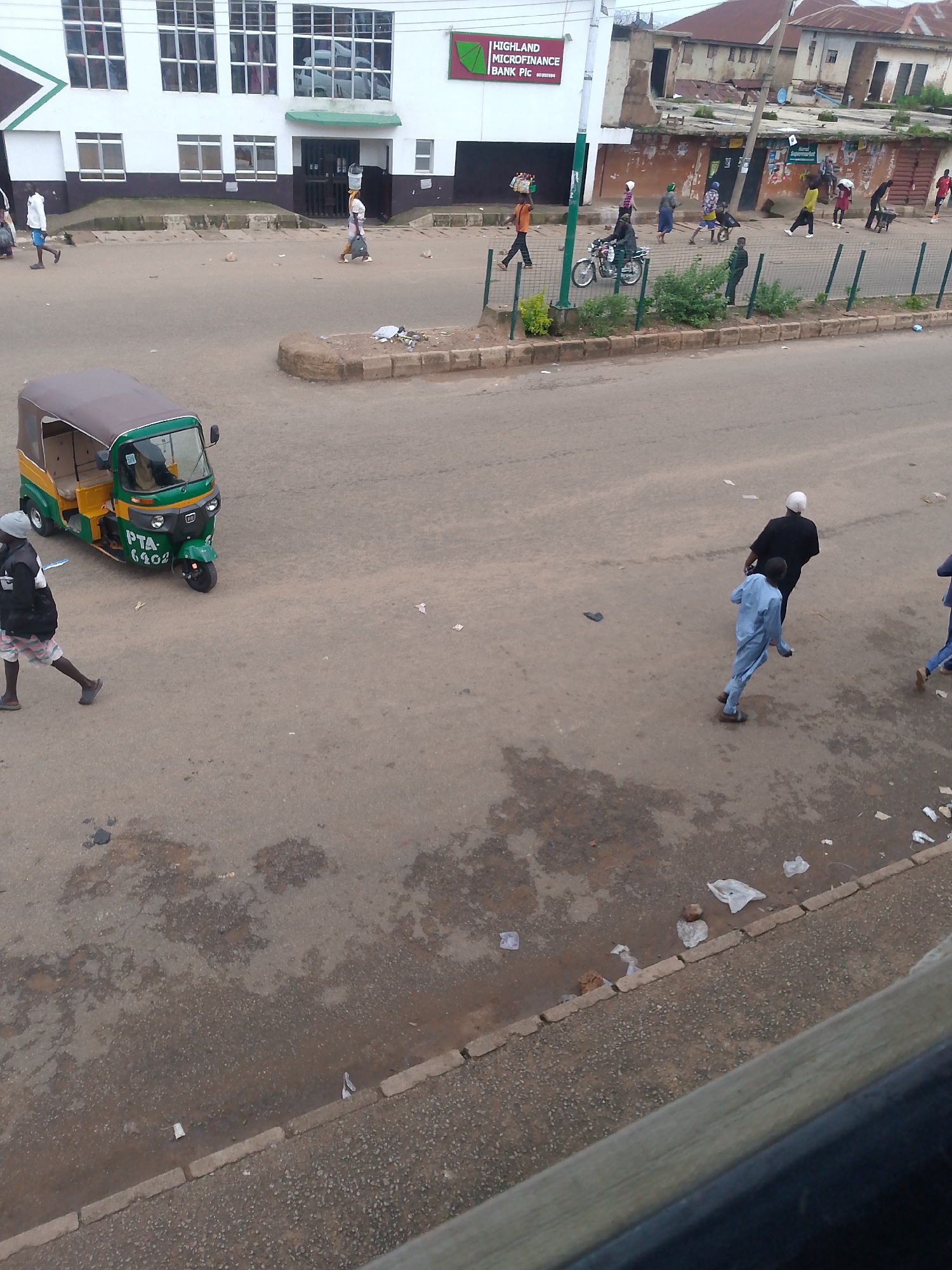 The image captures a street scene in Bukuru, Nigeria. In the foreground, a green and yellow auto-rickshaw is parked on the asphalt road. To its left, a person wearing a black jacket and patterned shorts walks away from the camera. Further up the road, a group of people are walking and interacting. Some are carrying items on their heads, while others are pushing carts. A motorcycle is also visible on the road. In the background, buildings line the street, including a white building with "HIGHLAND MICROFINANCE BANK PLC" signage. The ground is a mix of asphalt and dirt, with scattered debris. The sky is overcast, suggesting a cloudy day.
