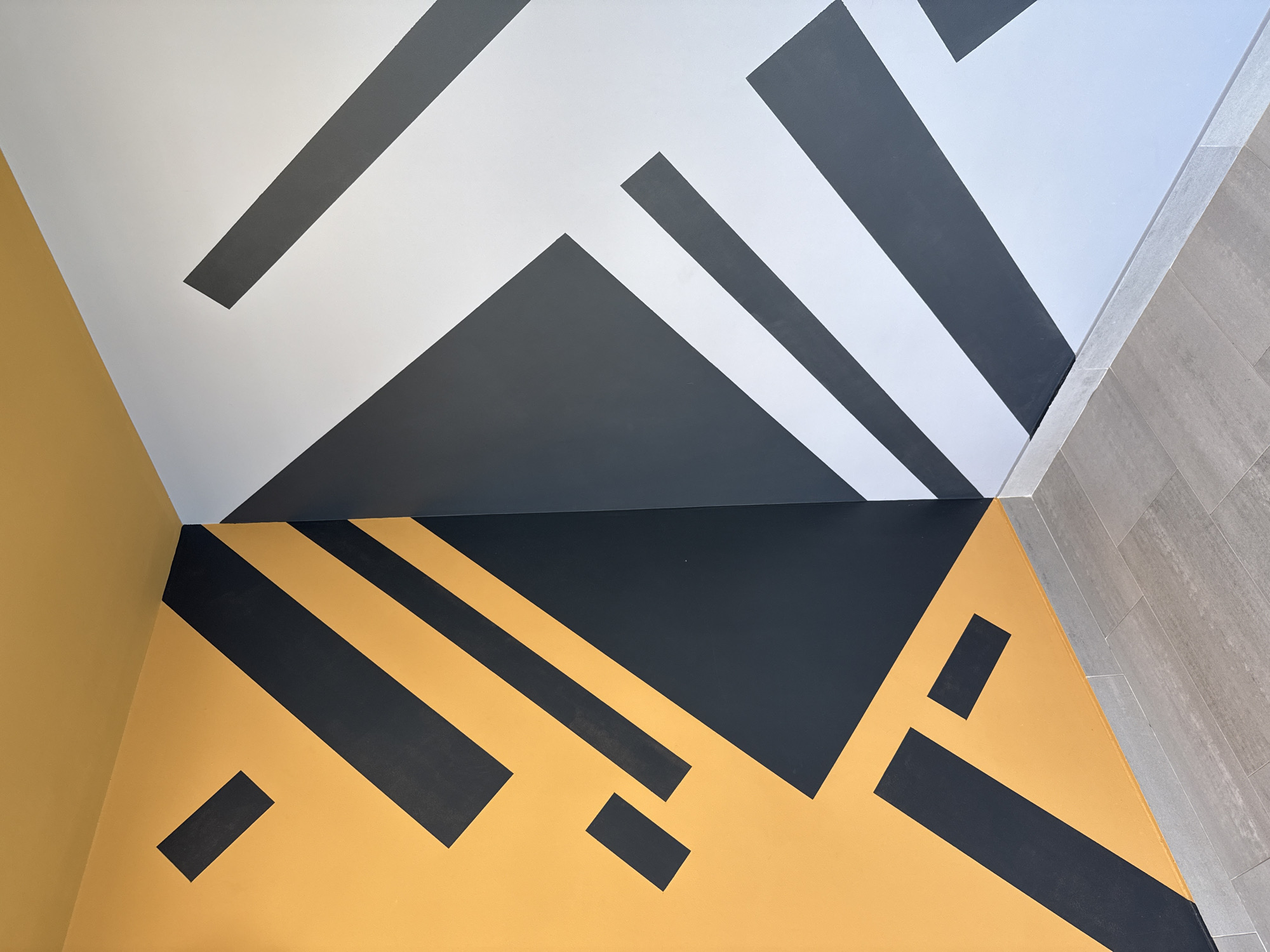 The image depicts a corner of a room. The wall on the left is painted mustard yellow. The wall in the back is painted white and gray. The floor is tiled gray. The walls and ceiling are decorated with a geometric pattern of black lines and shapes. There are no people visible. The location is unspecified and does not include any information about the city of Votkinsk, Russia.