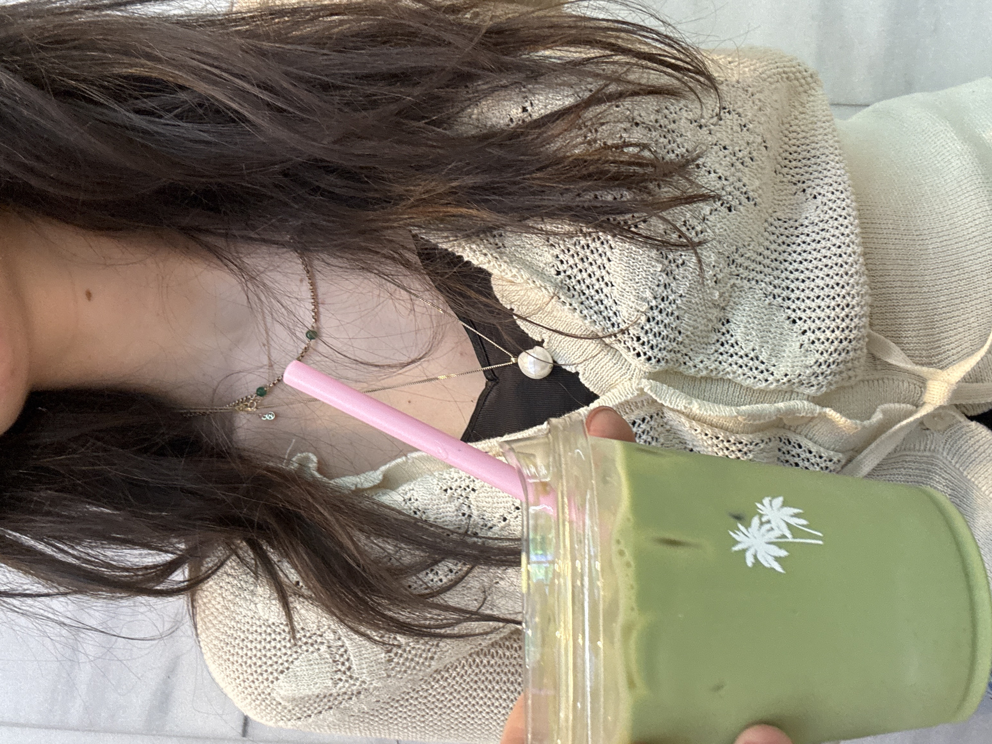 The image presents a close-up, top-down view of a person lying down or reclining, holding a green beverage. The upper body, from the neck to the mid-torso, is visible, along with dark brown, long hair fanned out around the shoulders. The person is wearing a light-colored, possibly cream or beige, open-knit or crocheted cardigan over a dark, likely black, inner top. A delicate gold chain necklace with small green beads and a larger, off-white, spherical pendant is draped across the chest.
The primary object of focus is a clear plastic cup filled with a vibrant green, possibly matcha or a similar green-hued drink, topped with some foam. A bright pink straw protrudes from the cup's lid. The side of the green cup features a white logo depicting two stylized palm trees. The person's hand, holding the cup, is partially visible at the bottom right.
The setting appears casual and indoor, possibly a cafe or home, with soft, diffused lighting suggesting daytime. No specific location cues are visible within the image to confirm the "New York, United States of America" context, but it fits a typical urban leisure scene. There is no visible text in the image apart from the palm tree logo. The overall impression is one of relaxation and enjoying a drink.