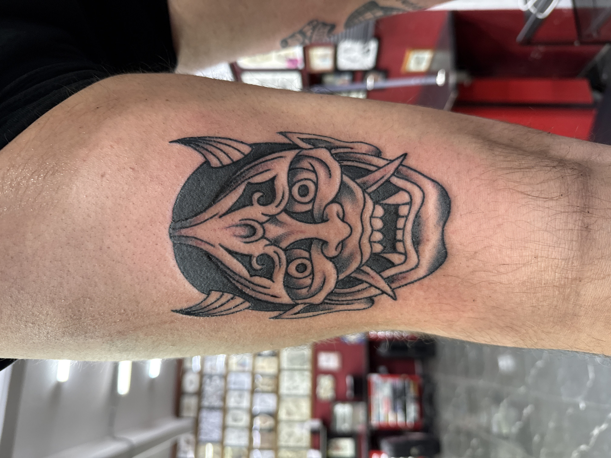 The image shows a close-up of a person's arm with a tattoo of a stylized demon mask. The tattoo is black and gray, featuring prominent horns, eyes, and teeth, set against a dark background. The arm appears to be lightly haired. In the background, there is a glimpse of a room, possibly a tattoo studio, with blurred items and a red toolbox, but its location in Moscow, Russia, cannot be determined from the provided image.