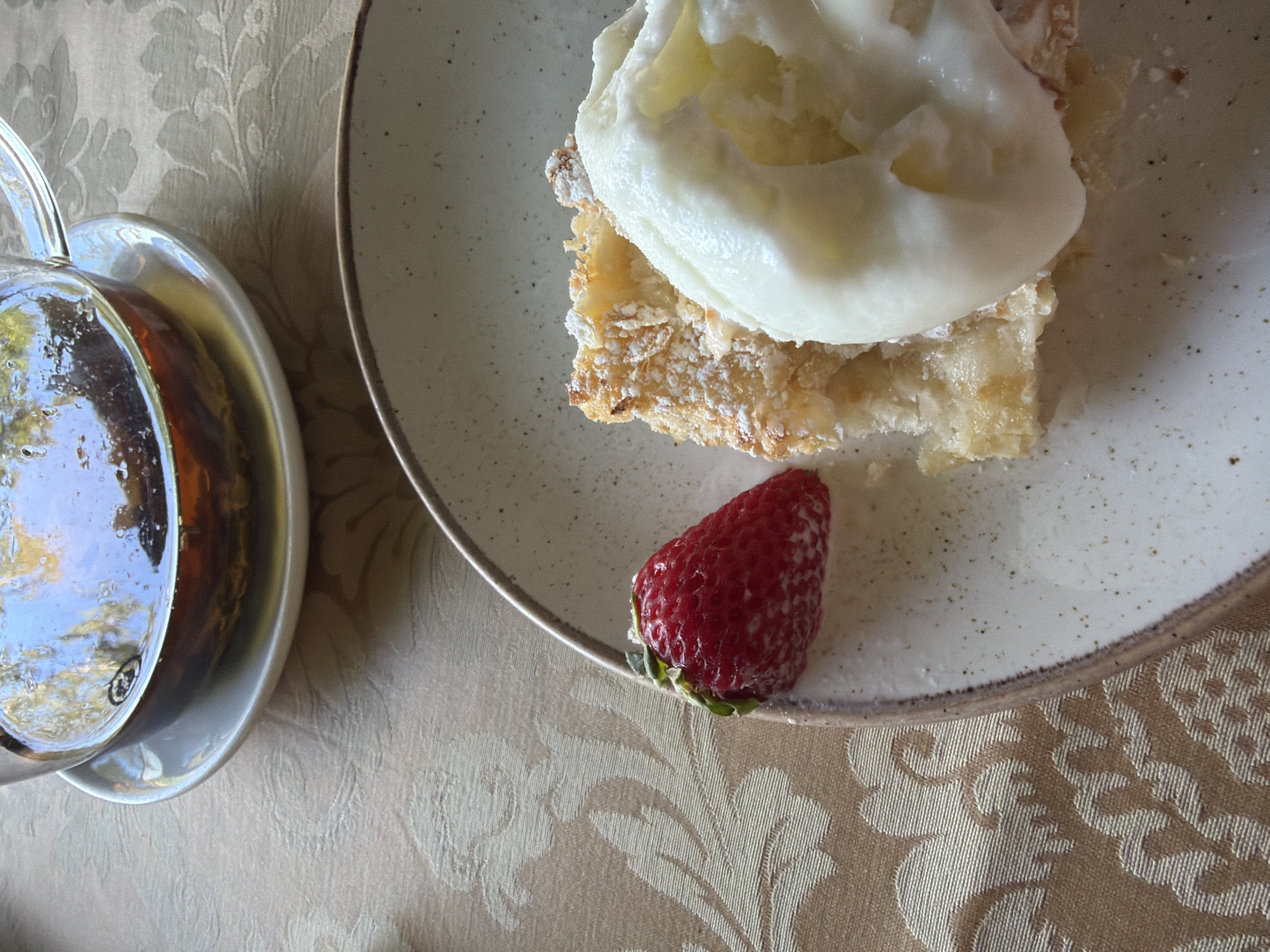 The image shows a close-up of a dessert served on a plate, next to a small glass teapot. The dessert appears to be a slice of pastry topped with a generous dollop of whipped cream or meringue, dusted with powdered sugar. A single, ripe strawberry sits to the side of the pastry. The plate is a light-colored ceramic with a speckled pattern. The teapot is made of glass and contains a golden-colored liquid, likely tea, with reflections of sunlight and possibly greenery on its surface. Both the plate and teapot rest on a table covered with a textured tablecloth featuring a floral pattern in shades of beige and cream. The scene suggests a moment of indulgence, perhaps an afternoon tea or a dessert break. There are no people visible, and the overall impression is one of quiet enjoyment. The lighting suggests it could be daytime, with natural light illuminating the scene. The location context provided is Svetlogorsk, Russia.