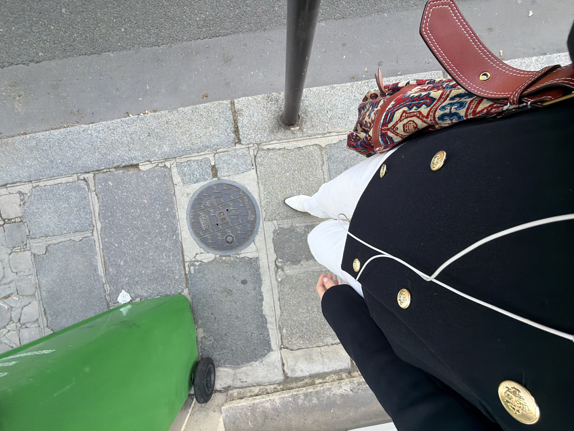 An overhead, first-person perspective photograph captures a person standing on a Parisian sidewalk adjacent to a street. The individual is wearing a black double-breasted coat adorned with large gold buttons, white pants, and white footwear. A white earbud wire is visible across the front of the coat. A brown leather shoulder bag with white contrast stitching and a multi-colored patterned textile insert, featuring red, blue, and beige geometric and floral motifs, is visible on the person's right side. The sidewalk is paved with irregular grey stone blocks. Embedded in the sidewalk is a round, metallic manhole cover with the inscription "GAZ" and additional partial letters visible, indicating Gaz de France, typical for Paris. To the left, a tall, bright green cylindrical public trash bin with a single black wheel at its base is positioned. To the right, a darker asphalt road surface is visible, separated from the sidewalk by a grey concrete curb. A dark metal pole extends vertically from the sidewalk edge on the right. Small pieces of debris, including a crumpled white item, are visible on the ground near the trash bin.