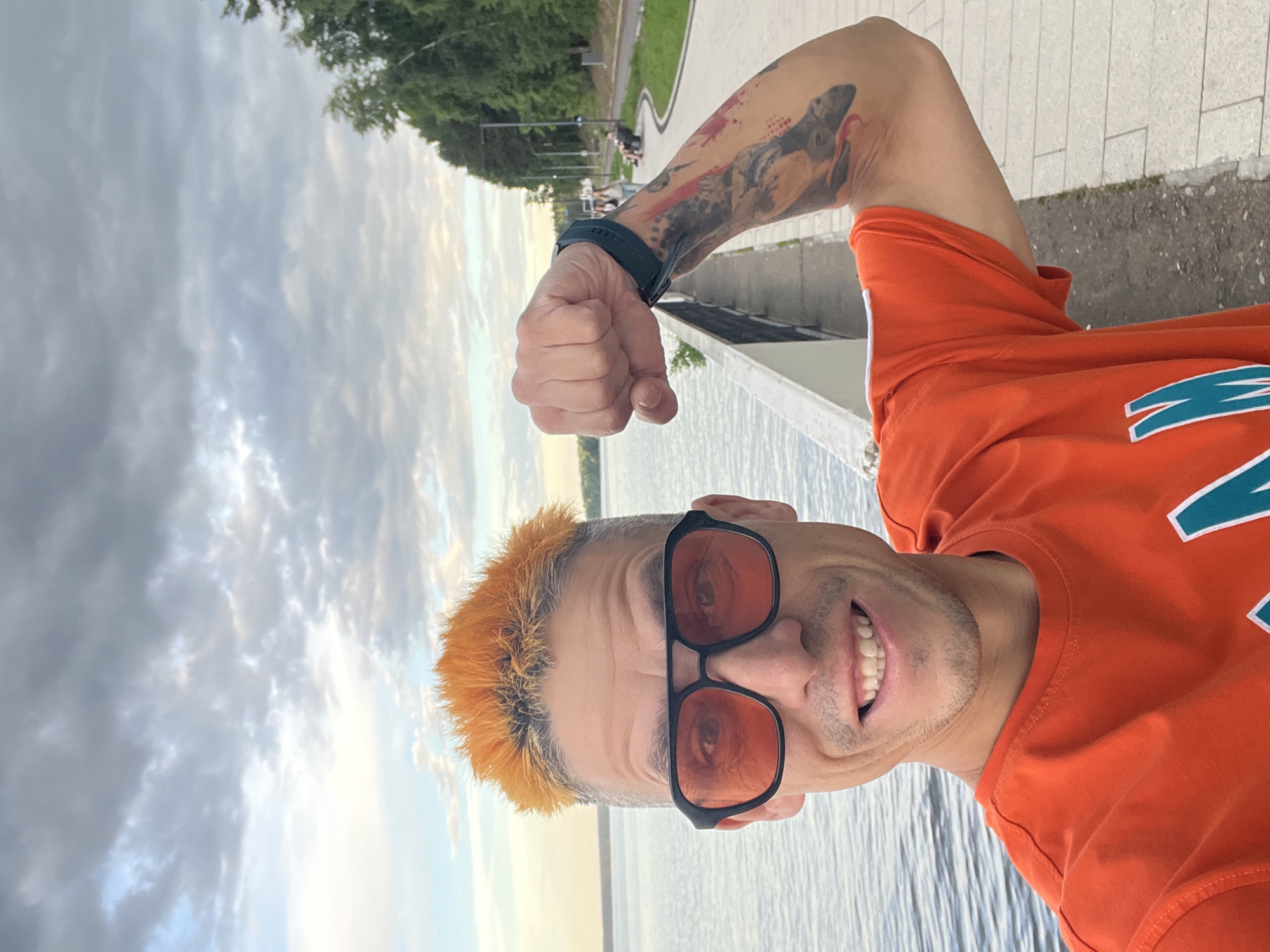 A man with brightly dyed orange hair and orange-tinted sunglasses is captured in a close-up, low-angle selfie. He is wearing an orange t-shirt with a teal and white graphic logo on the right side of his chest. His right arm is flexed, revealing a large, colorful tattoo on his bicep. He has a wide, genuine smile, showing his teeth. The background features a body of water, likely a river or lake, with a cloudy sky above. To the left, there is a paved walkway with trees and streetlights visible, suggesting an outdoor public area. The lighting indicates it is likely late afternoon or early evening. The scene is set in Chaikovsky, Russia.