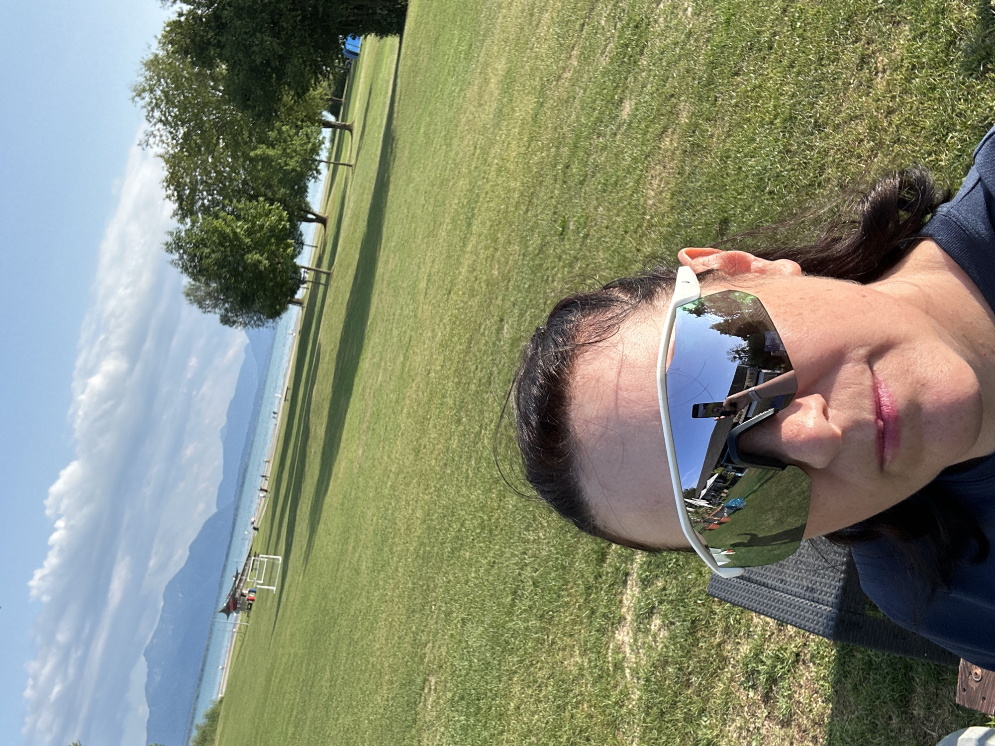 A woman with dark hair and dark eyebrows is taking a selfie outdoors, wearing white-rimmed sports sunglasses. The sunglasses reflect a scene of a lakeside area with mountains in the distance. The woman is sitting on a dark mesh chair, with a blue shirt visible. In the background, a green grassy field slopes towards a body of water, likely a lake, with mountains and a cloudy blue sky. Several trees are scattered across the field. A small structure with a red roof and people are visible near the water's edge. The reflection in her sunglasses also shows trees, a building, and what appears to be a phone being held up for the photo, along with her own reflection. The scene appears to be in Seeon-Seebruck, Germany, given the landscape.