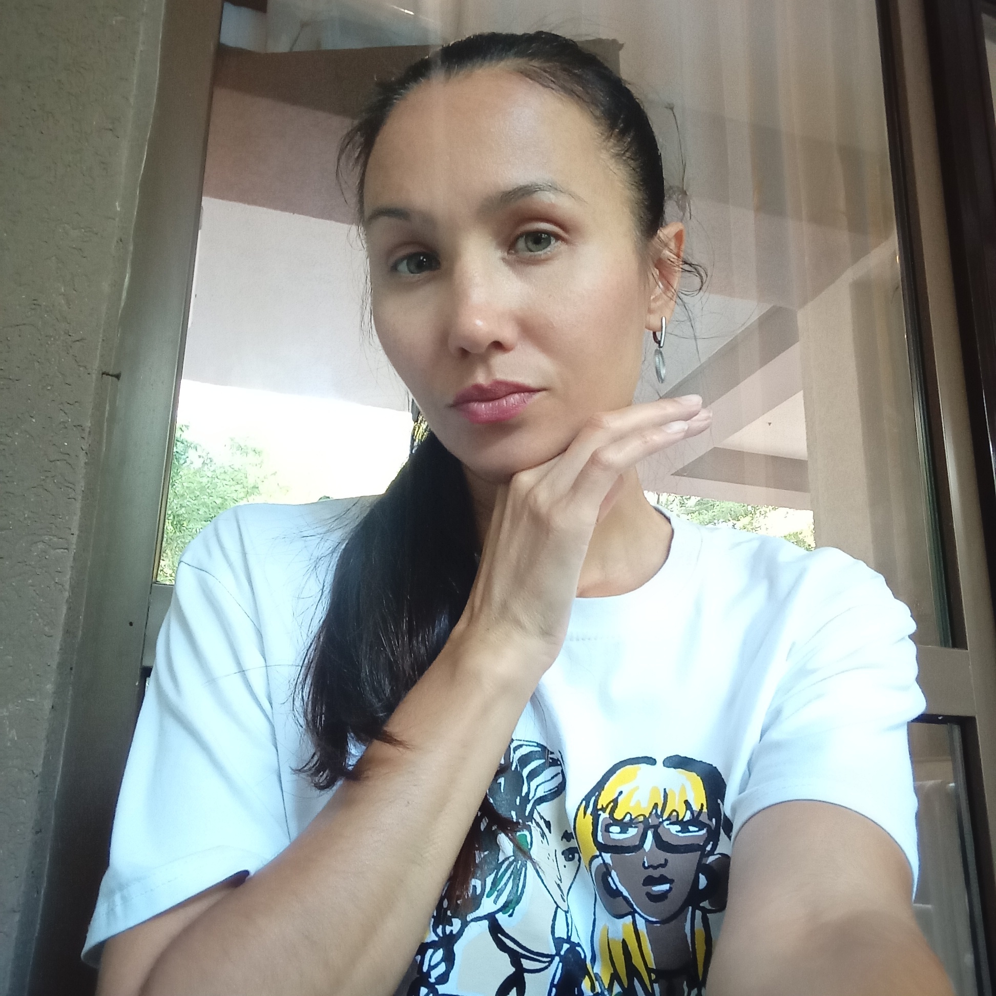 An outdoor, medium shot captures a woman in her early 30s posing for a selfie. She has long, dark hair pulled back into a ponytail, and she's wearing a white t-shirt with a graphic print of two stylized female faces. Her left hand is raised, touching her chin, and she's looking directly at the camera with a neutral expression. The background appears to be a balcony or patio area with a textured wall on the left and a glass door or window to the right, revealing greenery and part of a building's interior. The lighting suggests it's daytime. There is no specific indication of the city of Sochi, Russia, in the image.