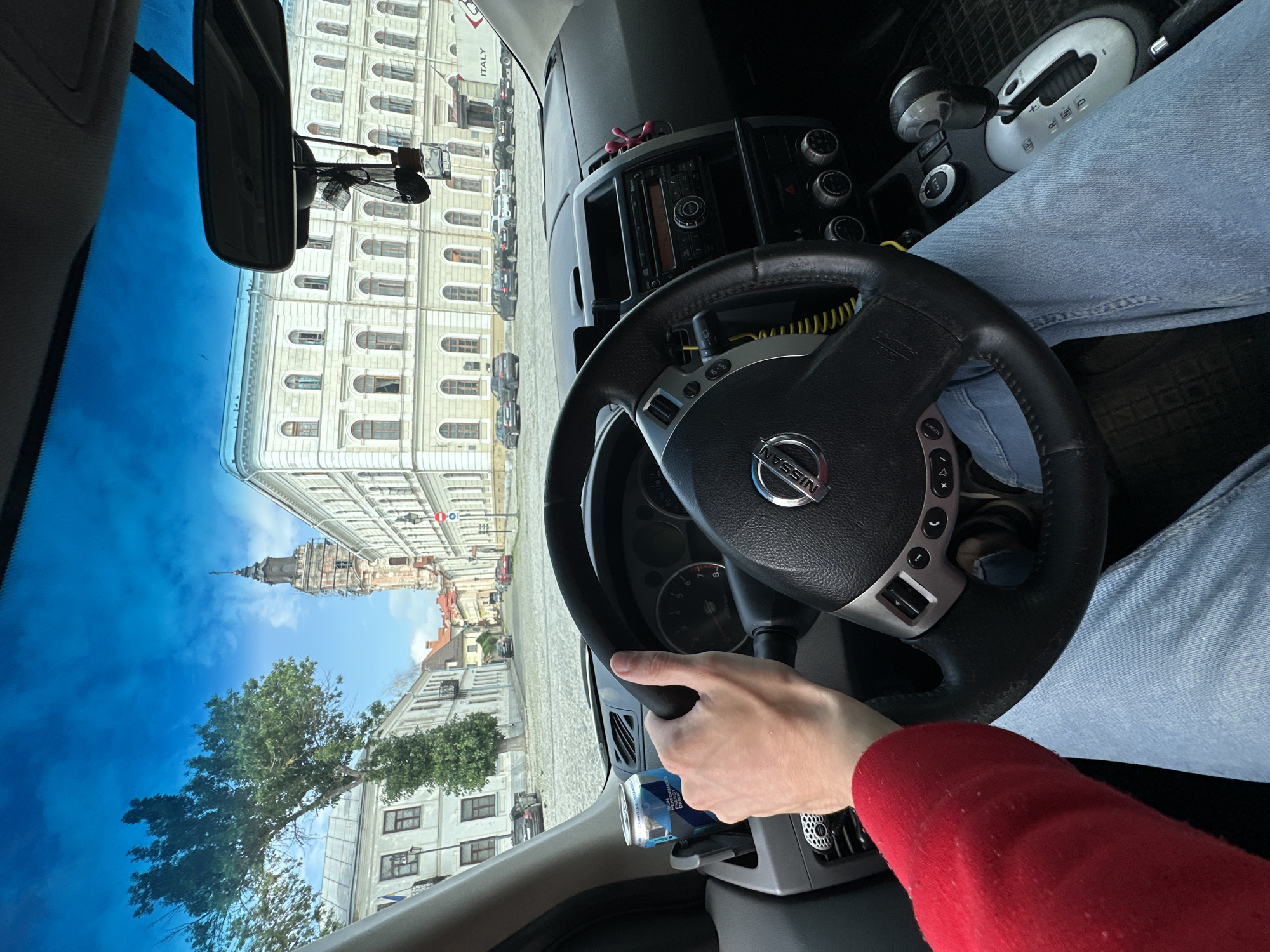 The image is taken from inside a Nissan car, looking out through the windshield. A person's hands are visible on the steering wheel, wearing a red sweater and blue jeans. The car is parked on a cobblestone street in what appears to be an old European city.
In the background, there are several buildings with classical architecture. One prominent building on the right is a large, multi-story structure with many arched windows and decorative elements. To the left of this building, a tall, ancient bell tower or church spire with a weathered facade and scaffolding is visible. The street is lined with parked cars, and trees with green foliage are scattered throughout the scene. The sky is blue with some scattered clouds.
Based on the architectural style and the presence of the bell tower and classical buildings, this scene is consistent with the city of Kamianets-Podilskyi, Ukraine, a city known for its historic Old Town and fortress. The specific location within Kamianets-Podilskyi is likely a square or street in the historic center, possibly near the Town Hall or other significant landmarks.