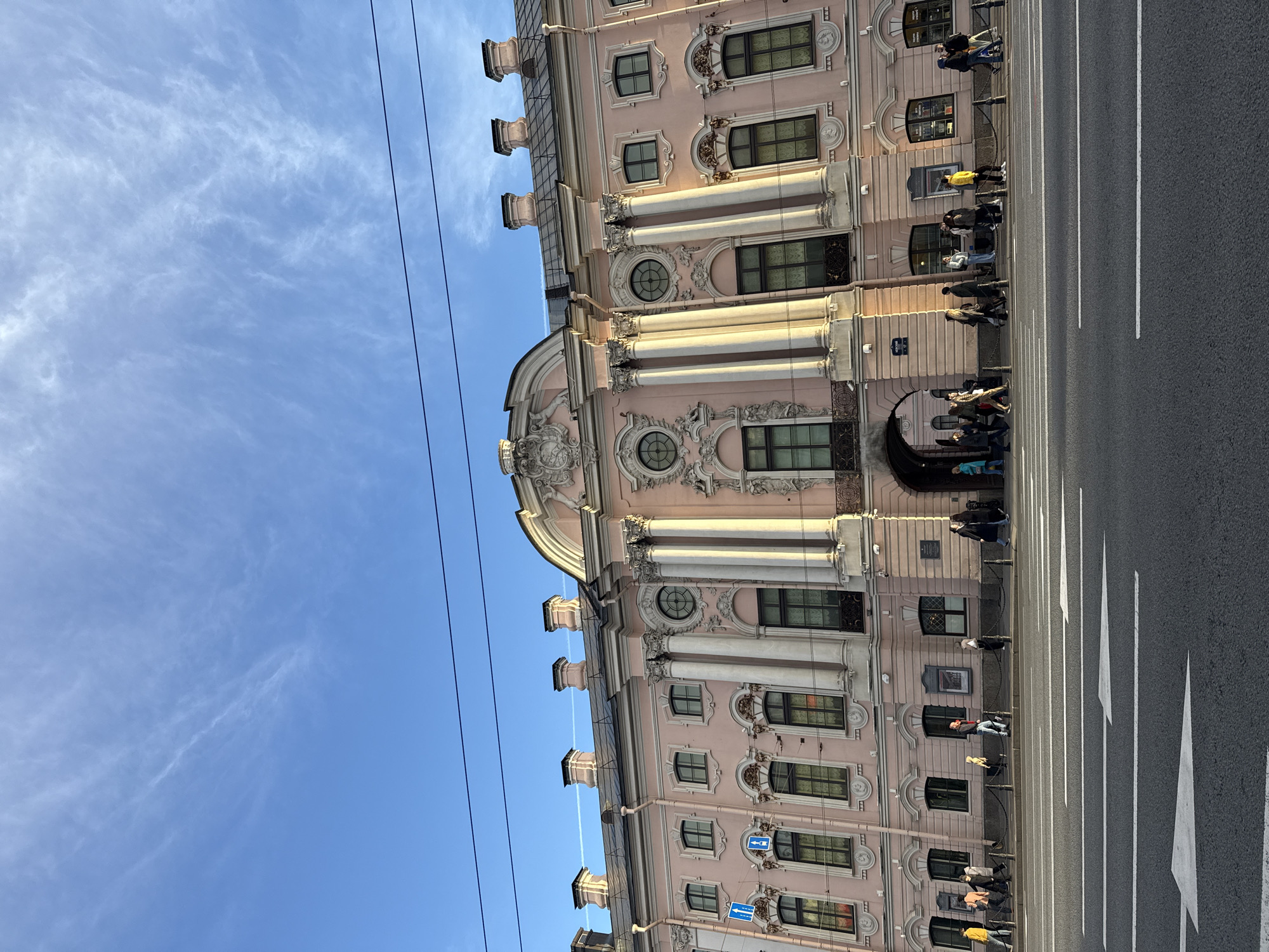 The image captures a vibrant street scene in Saint Petersburg, Russia, dominated by a grand, multi-storied pink building under a clear blue sky. The building exhibits ornate architectural details, including tall columns, decorative window frames (some rectangular, some circular), and sculpted embellishments, indicative of Baroque or Rococo style. Several small rooftop structures are visible along the cornice.
Along the base of the building, a sidewalk accommodates numerous pedestrians, casually walking and gathered near an arched entrance on the ground floor. People are dressed in various everyday attire. Adjacent to the sidewalk is a wide asphalt street featuring distinct white markings: dashed lines, solid lines, and prominent triangular arrows pointing in the direction of traffic flow.
The scene is bathed in bright daylight, suggesting a sunny late morning or afternoon, with a few wispy white clouds scattered across the sky. Dark electrical wires or cables traverse the upper left quadrant of the frame. Visible on the building's facade are a couple of blue rectangular signs; one clearly displays a white arrow pointing right, likely a traffic directive. Other smaller textual elements, such as a plaque near the arched entrance, are present but illegible. The image conveys the atmosphere of a bustling historic city street.