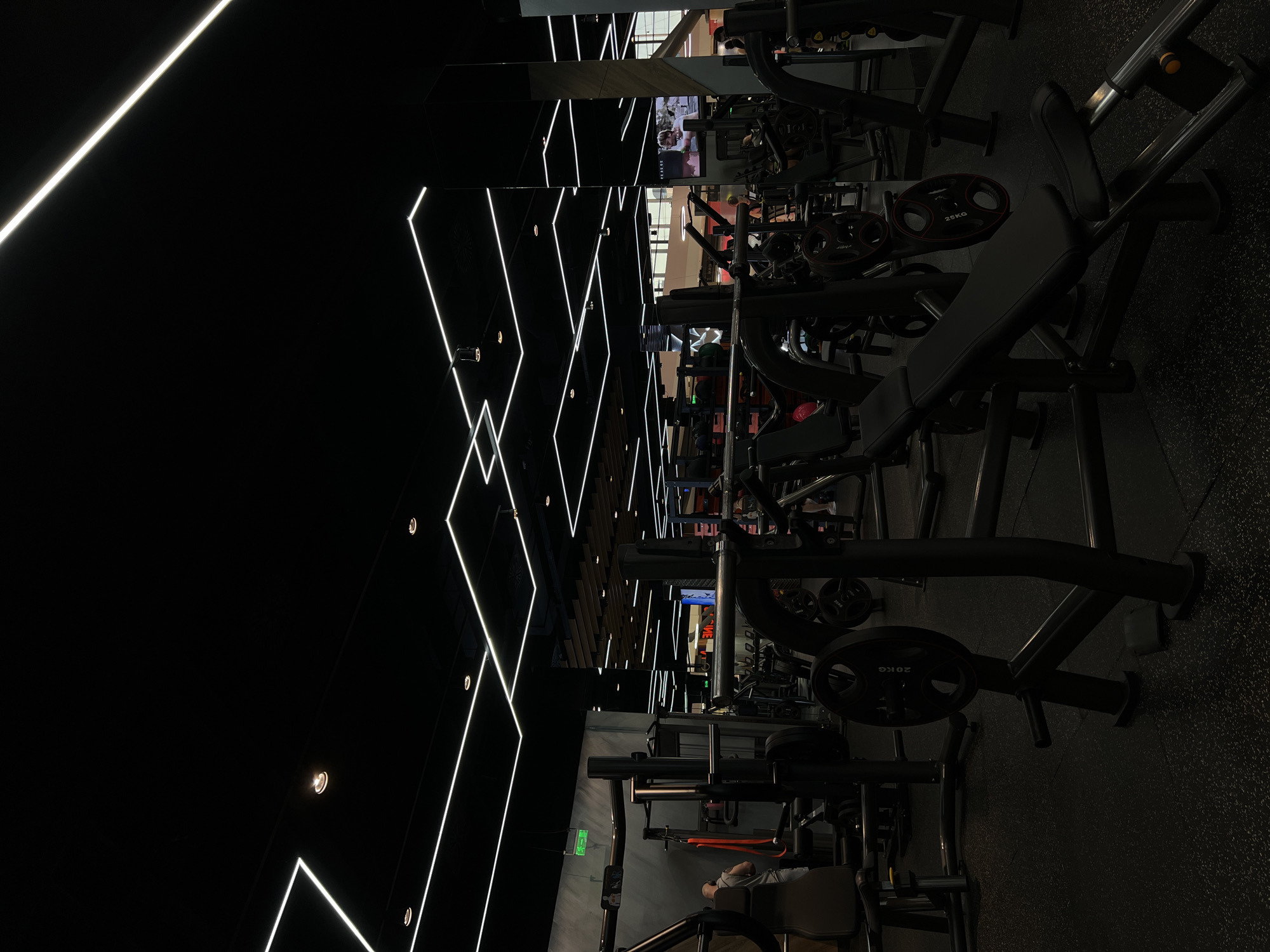 The image depicts the interior of a gym, likely in an urban setting but not discernibly Burgas. The gym contains various weight training machines, some of which have weight plates loaded onto them. The ceiling features geometric patterns created with bright white LED lighting. The floor is a dark, textured material, possibly rubber. Individuals appear in the background using the equipment, but their specific actions are not discernible due to the image's focus and lighting. The environment is dimly lit, emphasizing the artificial lighting and creating a modern aesthetic. The layout suggests a commercial gym space.
