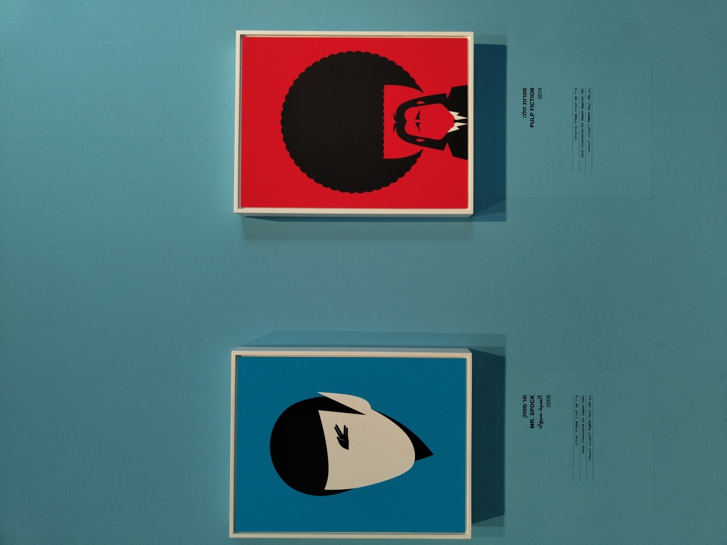 The image shows two framed artworks displayed on a teal-colored wall. The artwork on the left is a minimalist illustration of the character Mr. Spock from Star Trek, featuring his iconic black hair and pointed ears against a blue background. Below this artwork, text in Hebrew and English reads "MR. SPOCK" and "2009". The artwork on the right is a minimalist illustration of a character with a large Afro hairstyle and a red background, representing "PULP FICTION". Below this artwork, text in Hebrew and English reads "PULP FICTION" and "2014". Additional text below each artwork provides details about cover illustrations for Empire magazine. This scene is likely from an art exhibition or gallery, and given the Hebrew text, it is plausible that this location is in Tel-Aviv, Israel.