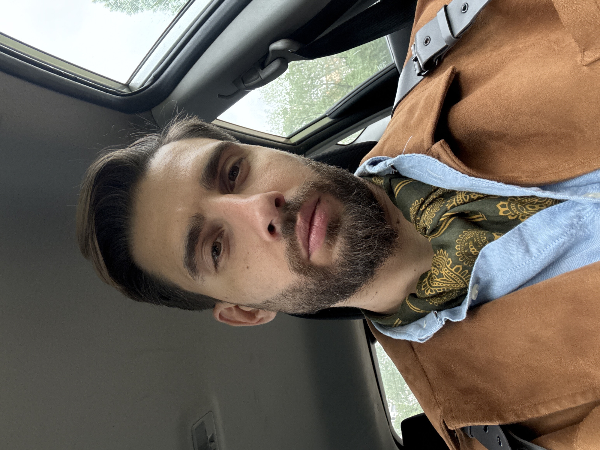 A man is positioned inside a car with an open sunroof. He has dark hair and a beard. He wears a blue shirt, a patterned green scarf, and a brown suede jacket with a black belt. The interior of the car is visible, including parts of the roof and the seat. Outside the sunroof, trees are visible. The location is unspecified. The man is looking into the camera.
