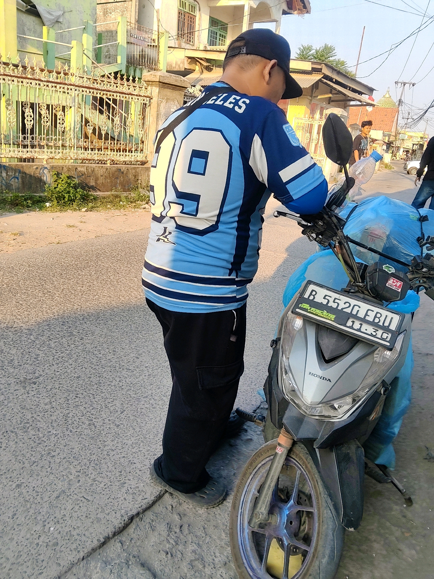 A person wearing a blue and white jersey with the number "9" prominently displayed on the back, a black baseball cap, and black cargo pants stands next to a silver Honda scooter. The scooter is partially covered in a bright blue tarp. The license plate of the scooter reads "B 5520 FBU". The person is looking down and appears to be interacting with the scooter or items on it. In the background, there are buildings with green and yellow facades, a metal fence, and overhead electrical wires, suggesting an urban or semi-urban environment. Sunlight casts shadows, indicating it is daytime. There are other people and vehicles visible in the distance, suggesting a street scene in a populated area. The overall scene depicts a person and their vehicle on a street. The location is identified as Mekarmukti, Indonesia.