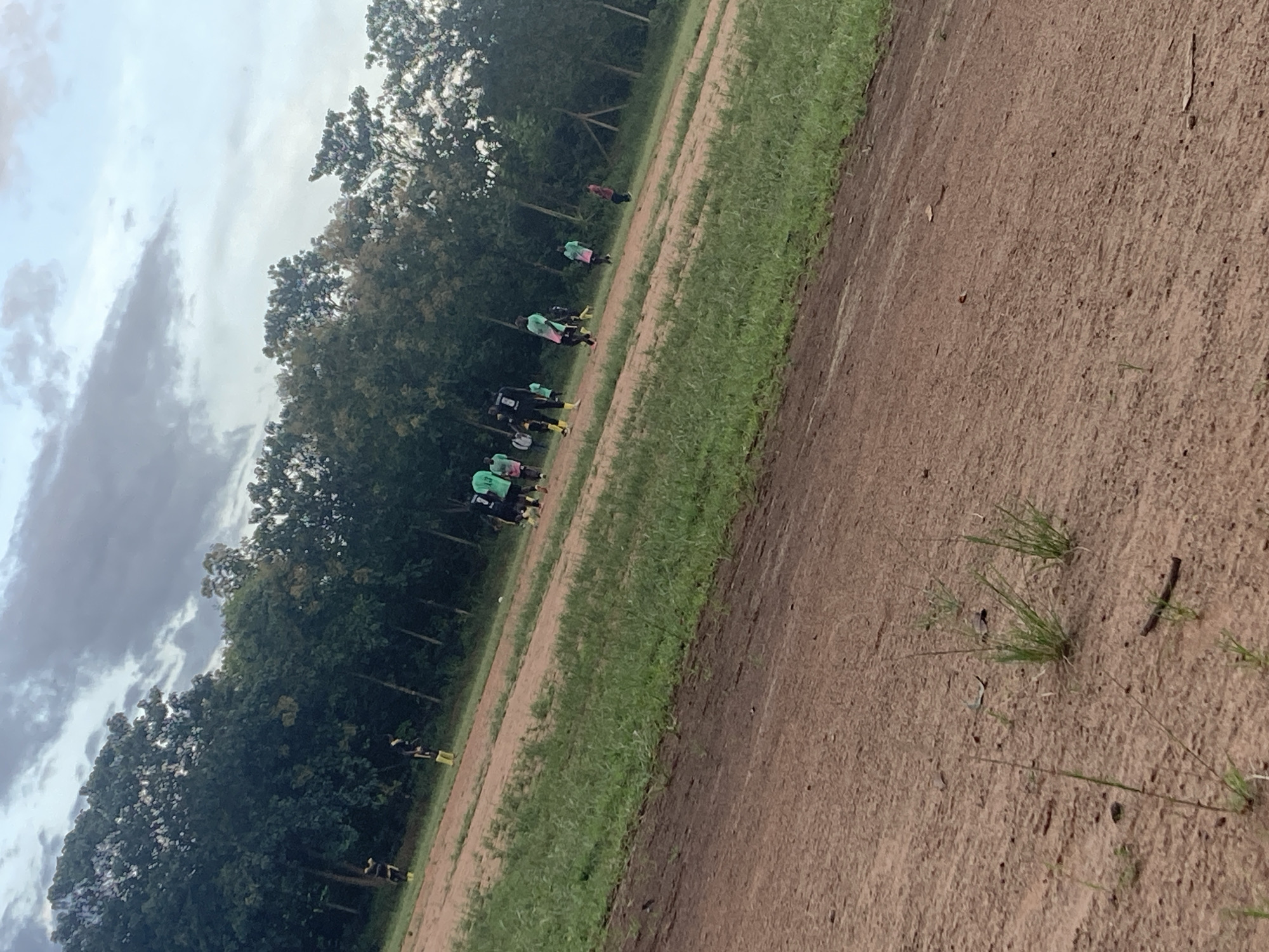 The image depicts a group of individuals, appearing to be teenagers or young adults, engaged in a soccer match on a dirt field in Miango, Nigeria. The field is characterized by its reddish-brown dirt surface, interspersed with patches of green grass. A line of trees forms a backdrop to the scene, with a cloudy sky overhead. The players are dressed in athletic attire, some in dark uniforms with yellow accents, and others in green jerseys. The game appears to be in progress, with the players scattered across the field, some in motion. The overall impression is one of a casual, outdoor sporting event in a rural or semi-rural setting.