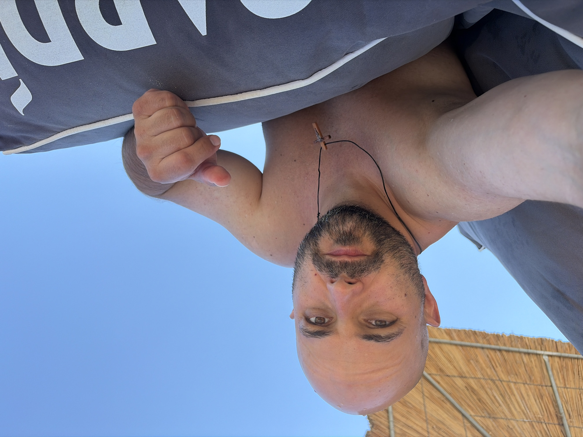 The image shows a bald, bearded man taking a selfie outdoors. He is wearing a necklace with a small wooden cross. The man is shirtless, and his skin appears wet, suggesting he may have been swimming. He is lying down on a gray cushion, and behind him, a thatched umbrella and a clear blue sky are visible. The scene appears to be a beach or a resort area. Based on the context provided in the prompt, the location is identified as the city of Kassandra Municipal Unit, Greece. The man is looking directly at the camera with a neutral expression. His right arm is bent, and his hand is resting on the cushion. The lighting suggests it is daytime, possibly midday, given the bright sunlight. The overall atmosphere is relaxed and summery.