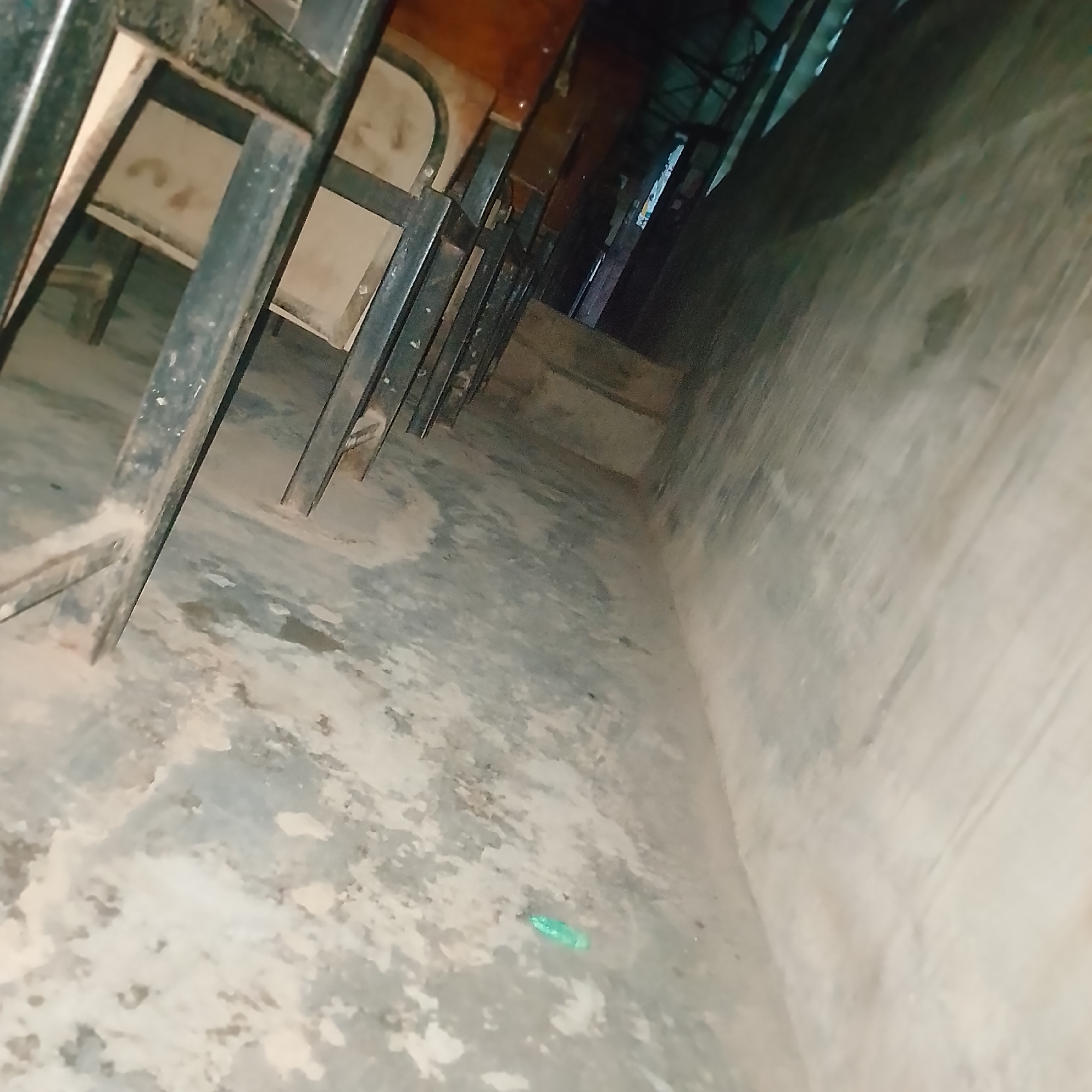 The image depicts a row of empty wooden chairs with metal frames, likely in a classroom or hall, at the Federal University of Agriculture, Abeokuta, Nigeria. The floor is a rough, concrete surface with some dirt and debris. The lighting is dim, with some natural light coming from an opening in the background, casting shadows on the floor and chairs. The overall impression is one of disuse and neglect.