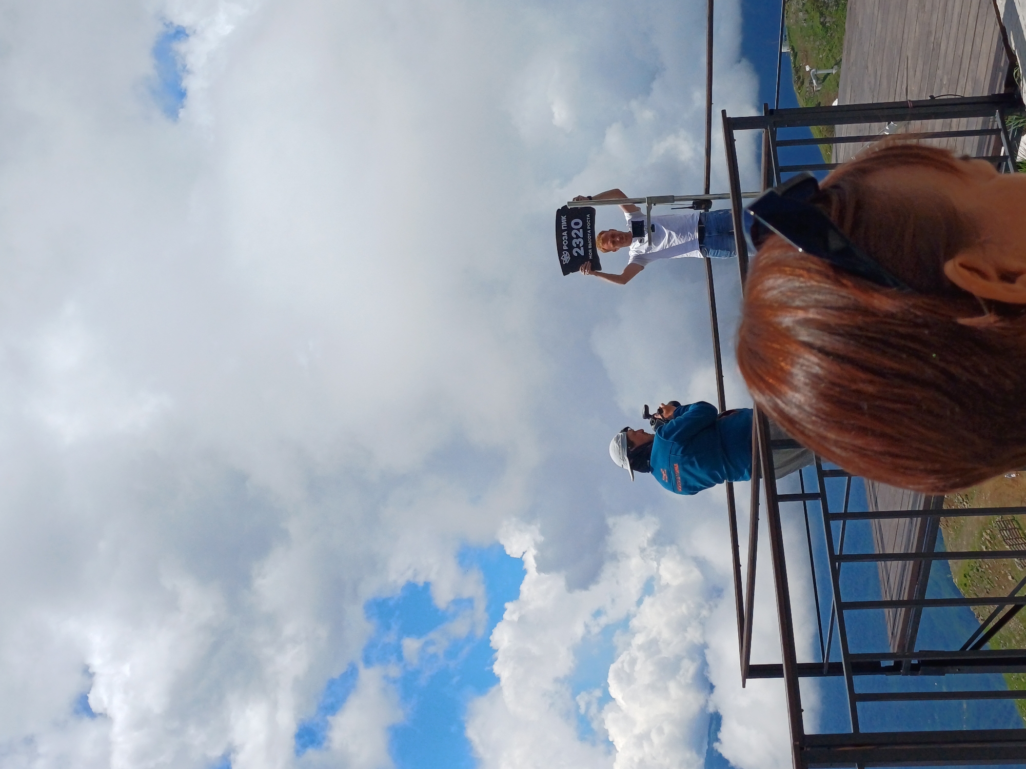 In the city of Estosadok, Russia, on a cloudy day with patches of blue sky, two individuals are visible on a mountain observation deck. One person, wearing a blue jacket and a white cap, stands facing away from the camera, holding a camera and appearing to be taking a photograph. The second person, a man with red hair wearing a white t-shirt and blue jeans, stands with his arms raised, holding a black sign with white lettering that reads "РОЗА ПИК 2320 МОЯ ВЫСОТА РОСТА". This sign indicates the location as Rosa Peak at an altitude of 2320 meters. The observation deck is made of metal railings and wooden planks. In the foreground, the back of a person's head with reddish-brown hair and dark sunglasses on top is visible, obstructing a full view of the scene. The background shows a mountainous landscape partially obscured by clouds.
