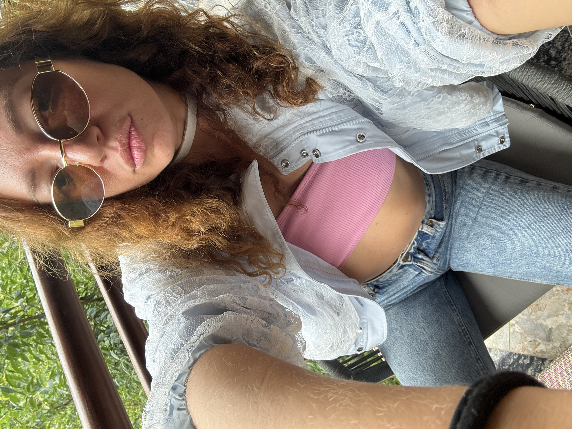 An indoor-outdoor selfie captures a woman with long, curly, reddish-brown hair looking directly at the camera with slightly pursed lips. She wears oval, gold-rimmed sunglasses, a narrow white choker, a pink ribbed bandeau top, and a light blue jacket with a white lace overlay and silver snap buttons, worn open. She is also wearing light blue denim jeans, seated in a dark grey woven chair. Her right forearm, showing light hair, is visible in the foreground. The background features a dark brown wooden railing and lush green foliage, suggesting a balcony or patio setting in Púng Luông, Vietnam. Light-colored, patterned tiles are visible on the floor.