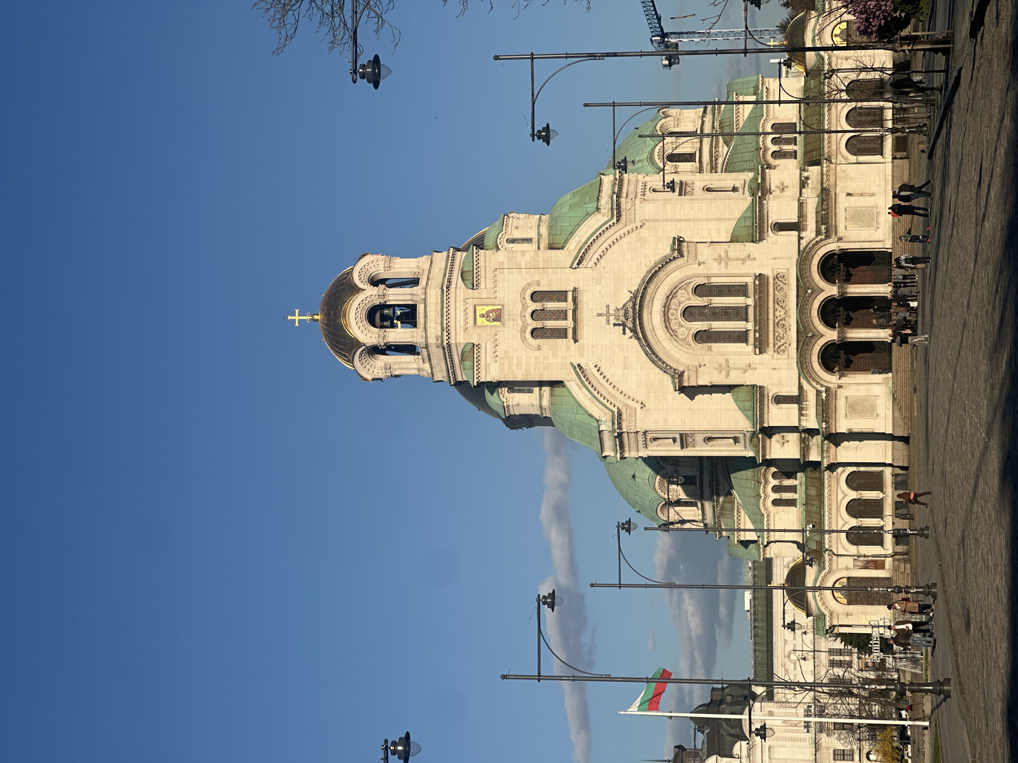 The image depicts the Alexander Nevsky Cathedral in Sofia, Bulgaria, viewed from a low angle. The cathedral is made of light-colored stone with green domes and a prominent bell tower topped with a golden cross. The Bulgarian flag is visible on a pole in the foreground. Several people are walking on a paved area adjacent to the building. Street lamps are positioned along the side of the cathedral, and a portion of a construction crane can be seen in the background. The sky is clear and blue.