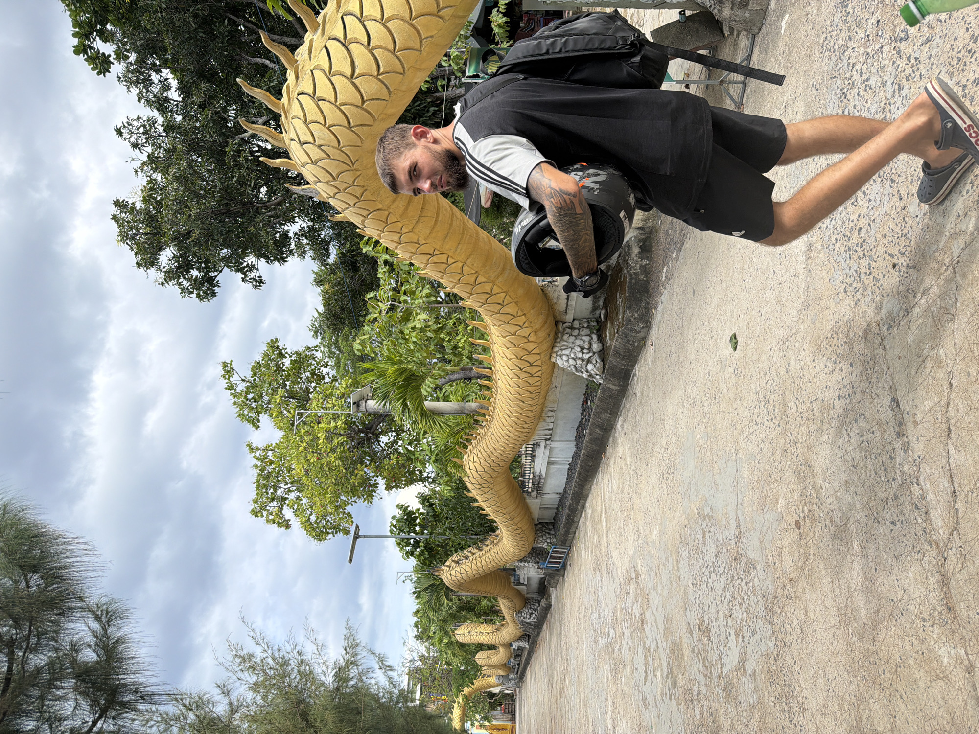 An outdoor, daytime shot in Cam Ranh, Vietnam, features a man standing on a concrete path next to a long, winding golden dragon statue. The image is taken from a slightly low angle, looking up towards the man and the dragon, with a cloudy sky and abundant green trees forming the background.
The man, with a beard and visible tattoos on his arms, is wearing a black t-shirt with grey and white striped sleeves, black shorts, and dark Crocs sandals. A black backpack is worn over his shoulders, and he holds a black motorcycle helmet with orange accents in his left hand. He is looking directly towards the camera with a neutral expression.
The prominent golden dragon statue snakes along the edge of the concrete path. Its body is covered in detailed scales, and it features a series of spines running down its back. In the distance, more segments of the dragon statue are visible, suggesting it extends for a significant length.
The surroundings include lush green trees with dense foliage, some palm trees, and what appears to be utility poles or light fixtures. The concrete path shows some wear and texture. A partially visible green bottle is seen near the man's feet in one of the crops. The text "CROCS" is visible on the strap of his sandal. The overall scene suggests a cultural or tourist attraction, likely a temple or garden, in Vietnam.
