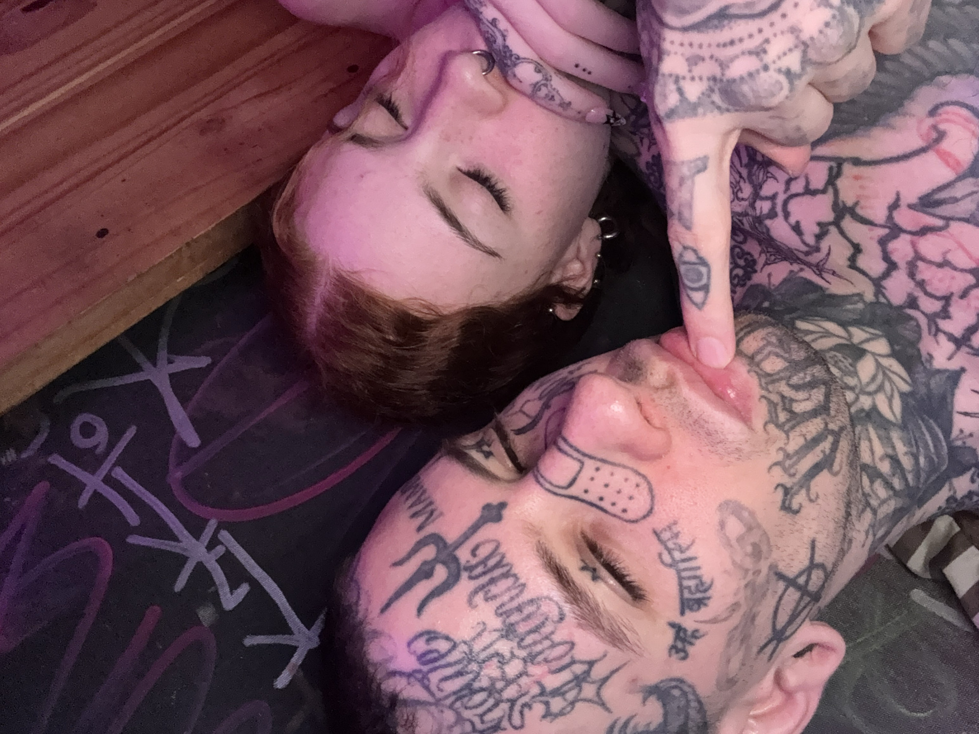Two people are lying down, close together. The person on the right side of the frame has extensive tattoos on their face and body. Their index finger is touching their upper lip. The other person has red hair and multiple ear piercings. They are lying on a surface with graffiti. Part of a wooden structure is also visible. The location is not discernible from the image.