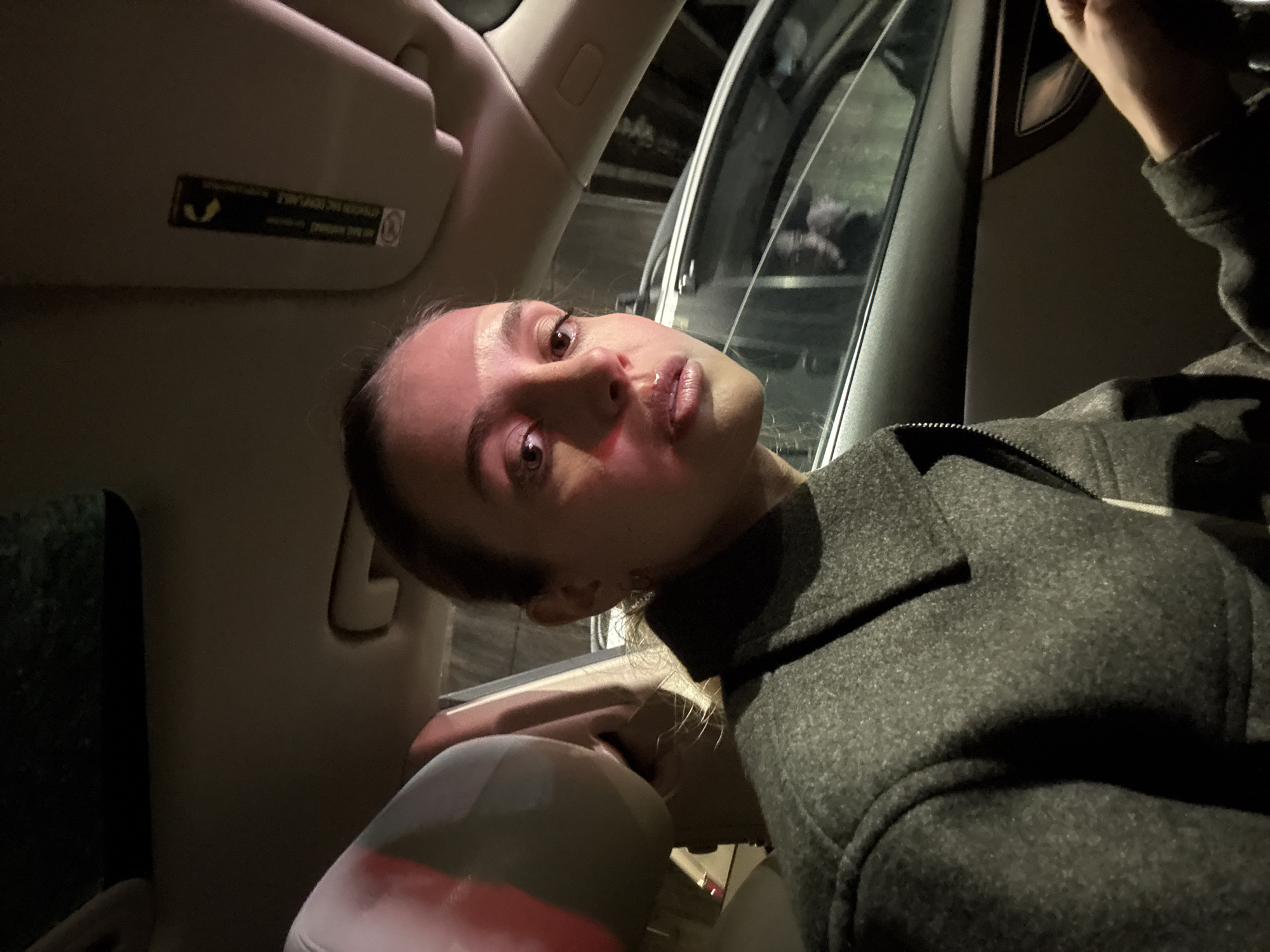 A person is sitting in the passenger seat of a car. The car's interior is visible, with the sun visor and door frame in the background. The person is wearing a grey coat. Outside the car window, a blurred urban environment is visible. It is nighttime. Without any additional clues, the location cannot be determined with sufficient precision to confirm that it is Tbilisi, Georgia.