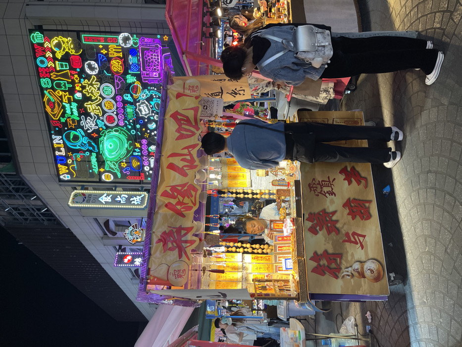 The scene is a street stall in Hang Zhou Shi, China, located at 30.264 latitude and 120.156 longitude. The stall is brightly lit and decorated with colorful lanterns and signs with Chinese writing. The stall is selling various trinkets and souvenirs, including small, brightly colored figures. There are three people in the scene: a man behind the counter, a man in front of the counter, and a woman standing to the right. The man behind the counter is sitting and appears to be selling something to the man in front of the counter. The woman is standing with her back to the camera, observing the scene. The environment is crowded, with other stalls and people in the background. The buildings in the background are illuminated with brightly colored neon signs. The scene is dynamic and bustling with activity.
