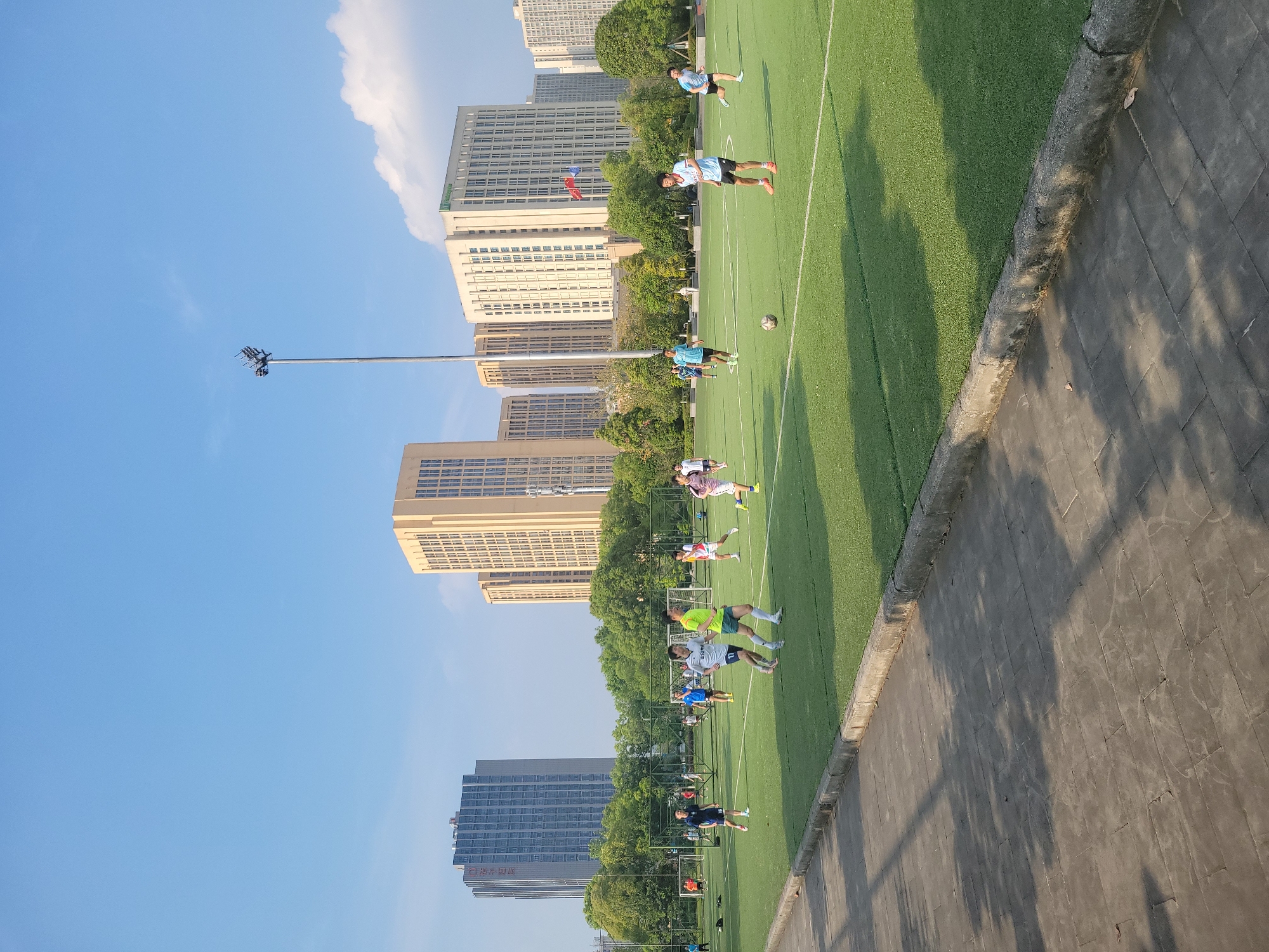 A horizontal outdoor shot depicts a soccer field in Chongqing High-tech Zone, China. Several people are practicing on the field. Buildings and trees line the field's perimeter. A tall light pole stands to the left, reaching into a blue sky with a few clouds. The field is bordered by a sidewalk with shadow patterns. A soccer ball is visible on the field.