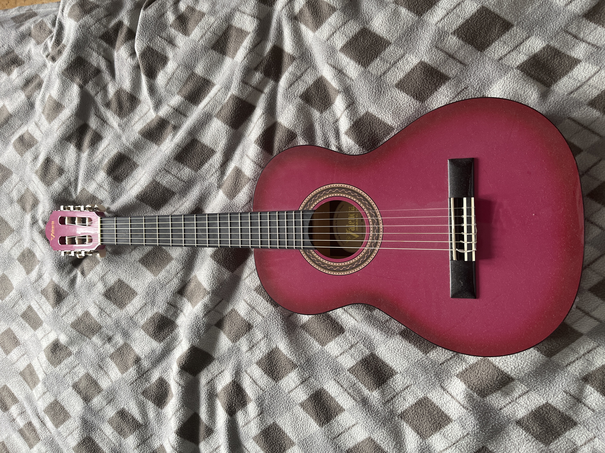 A pink guitar lies on a grey and brown checkered blanket. The guitar is positioned vertically, with the headstock at the top and the body at the bottom. The blanket is textured with a repeating diamond pattern. The lighting suggests an indoor setting, possibly a room with natural light. There are no people or any other objects visible in the image. The scene is static and appears to be a still life. There is no information to suggest this image is from Moscow, Russia.