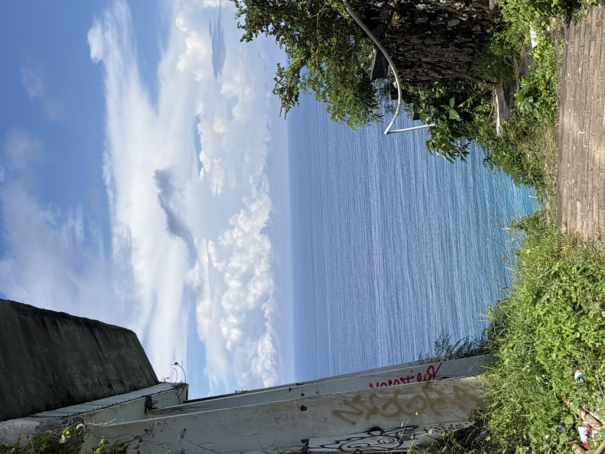 From Pecatu, Indonesia, this outdoor photograph features a view of the ocean and sky from an elevated perspective. The ocean occupies the central portion of the scene, reflecting the blue sky. Above it, the sky is partly covered with white, puffy clouds. On the left, the edge of a dark-colored building with graffiti along a white concrete wall is visible. On the right, green vegetation grows along the edge of a stone pathway. A metal pole is visible among the vegetation. The overall impression is a natural landscape juxtaposed with urban elements.