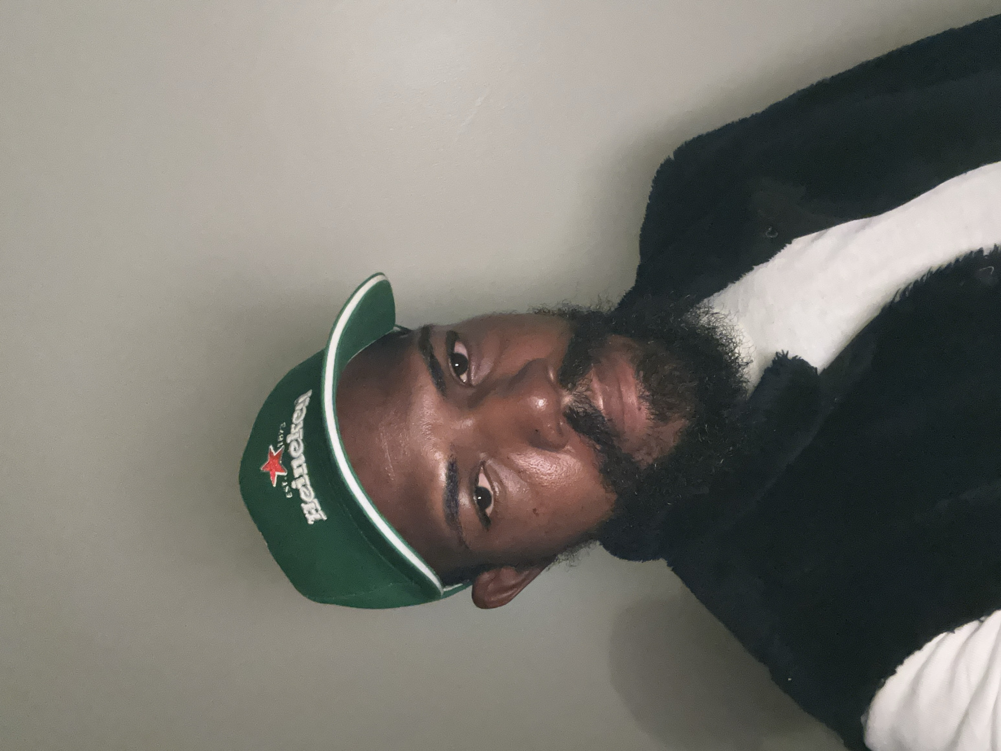 A man wearing a green Heineken baseball cap and a black jacket over a white shirt is captured in a close-up, waist-up portrait. The man has dark skin, a beard, and is looking towards the camera. The background is a plain, light-colored wall. The image appears to be taken indoors, possibly in a residential setting. The lighting is somewhat dim, casting subtle shadows. The overall mood of the image is casual. There is no information about the location in Lagos, Nigeria, within the image itself or its metadata.