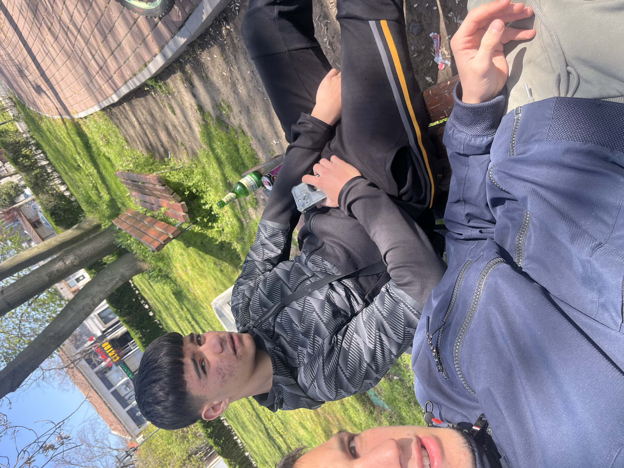 The image depicts two young men sitting on a park bench in Saedinenie, Bulgaria. One of the men is wearing a gray and black patterned hoodie and black pants with a yellow and gray stripe. He is holding a phone in his left hand. A green glass bottle is visible on the bench next to him. The second man is partially visible, wearing a navy blue jacket. The bench is located in a grassy area near a sidewalk and a brick-paved area. Buildings and trees are visible in the background under a partly cloudy sky.
