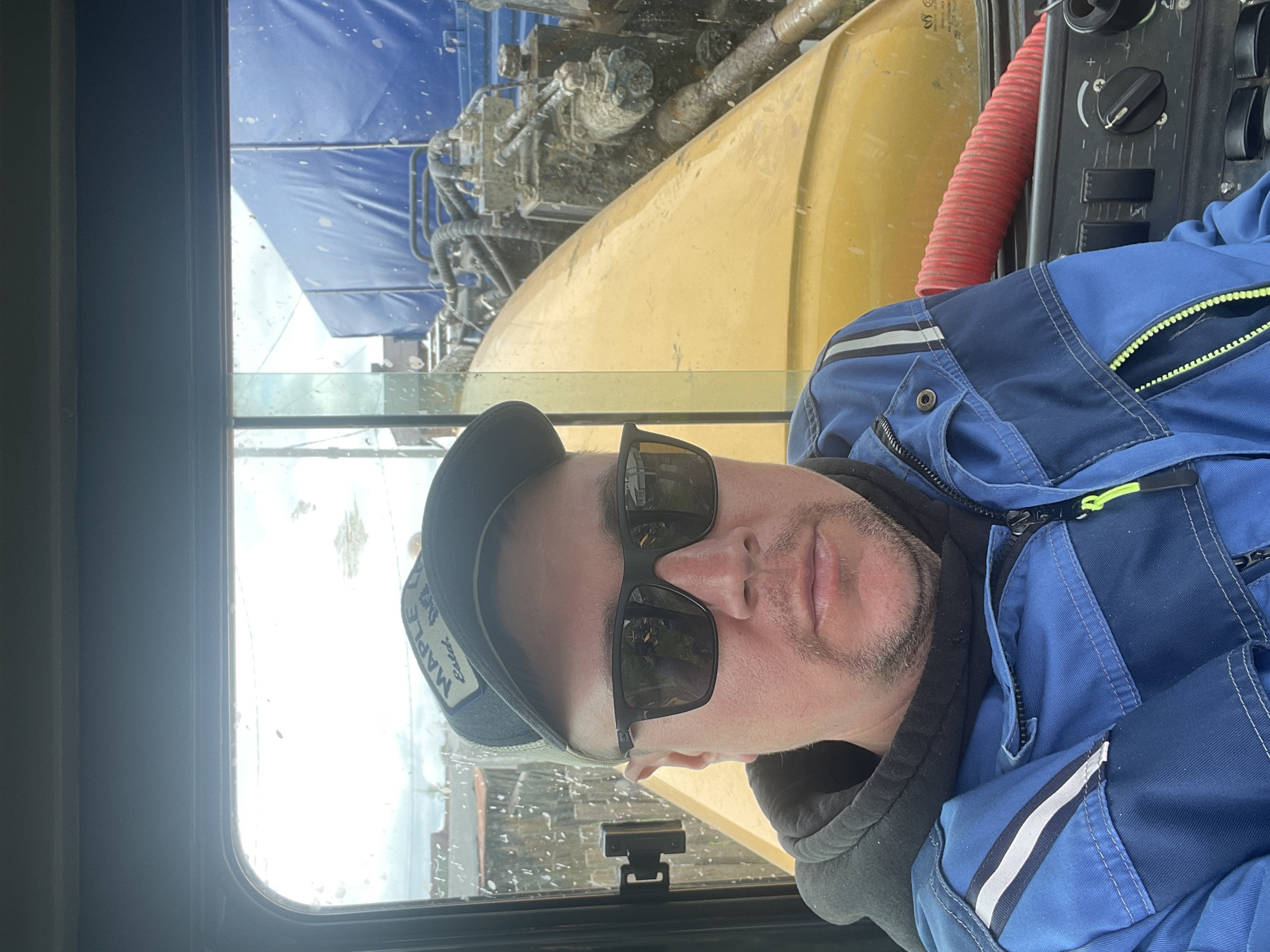 A male subject is depicted in a selfie taken from inside the cab of heavy machinery in the city of Железнодорожный, Russia. The man, appearing middle-aged with stubble, wears dark sunglasses, a black baseball cap with a "MAPLE Estd." patch, a black hoodie, and a blue work jacket with white and black reflective stripes on the left shoulder and a neon yellow zipper pull. He is looking directly at the camera. The interior of the vehicle's cab frames the scene, with a dark gray overhead panel and window frame visible. The windshield is splattered with water drops and dirt.
Through the windshield, the exterior environment is observed. Immediately in front of the man and to his right, the yellow bodywork of the heavy machinery is visible, heavily soiled with mud and grime. Beyond this yellow structure, intricate mechanical components, including hydraulic hoses, pumps, and valves, are exposed and covered in a thick layer of dirt. Further in the background, a large blue tarpaulin covers a load on what appears to be a truck or trailer, also splattered with dirt. The sky beyond is overcast. On the lower right side of the cab interior, a control panel with various switches and a rotary dial is partially visible, along with a red corrugated tube.