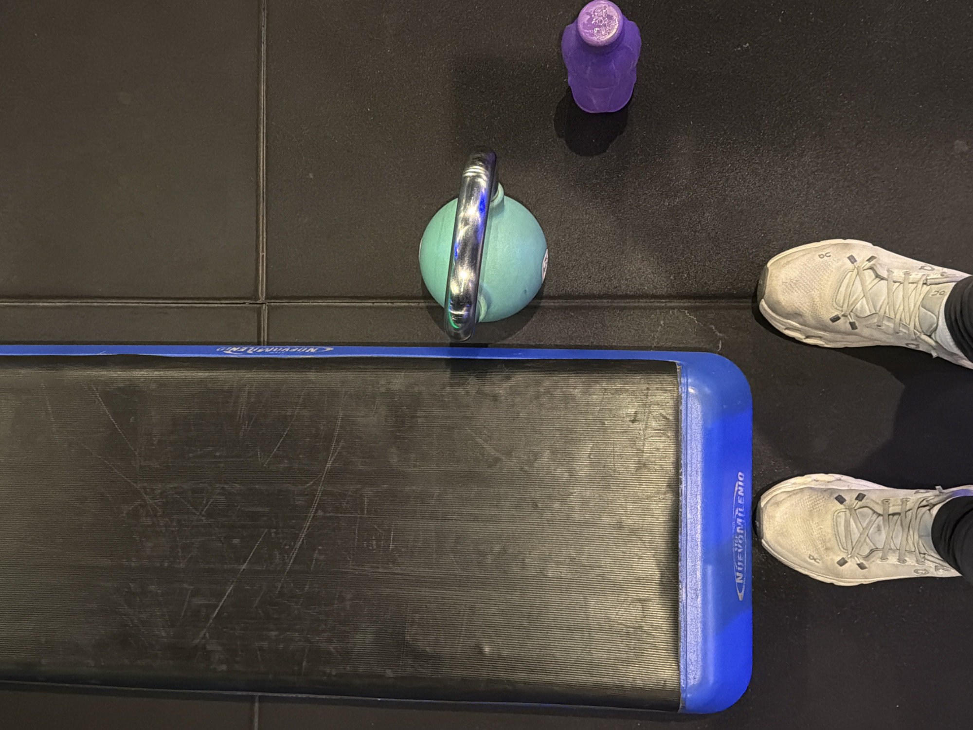 The image depicts a gym setting. A black aerobic step with blue edging and the word "NUEVOMILENIO" is visible on one side. Adjacent to the step is a teal kettlebell with a chrome handle. A purple water bottle is positioned near the kettlebell. To the right of these objects are a person's feet wearing white athletic shoes with black leggings. The floor appears to be composed of black tiles.
There is no information to confirm the location is in the city of Buenos Aires, Argentina.