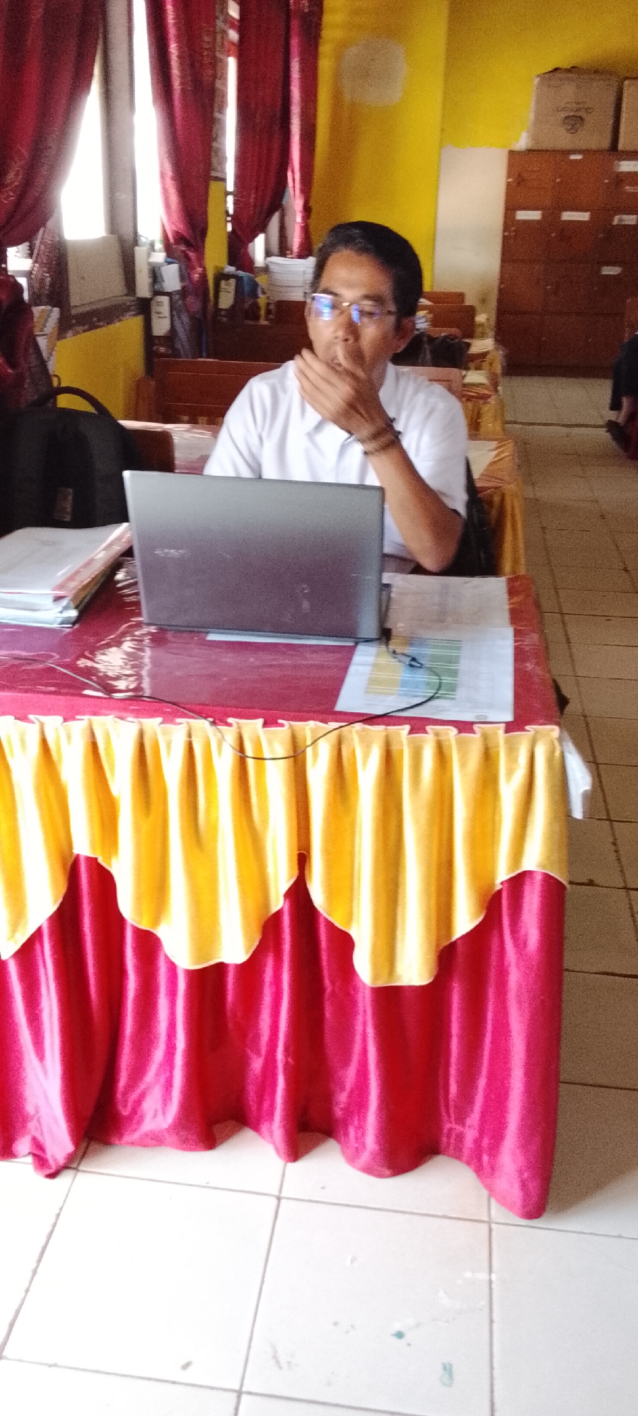 A man wearing glasses and a white collared shirt is seated at a table in what appears to be a classroom or meeting room. He is looking down at a laptop computer in front of him, with his hand covering his mouth. The table is draped with a red cloth that has a yellow valance along the front. Behind the man, there are shelves filled with boxes and papers, and red curtains hang on the wall. The floor is tiled. The scene suggests an educational or professional setting.