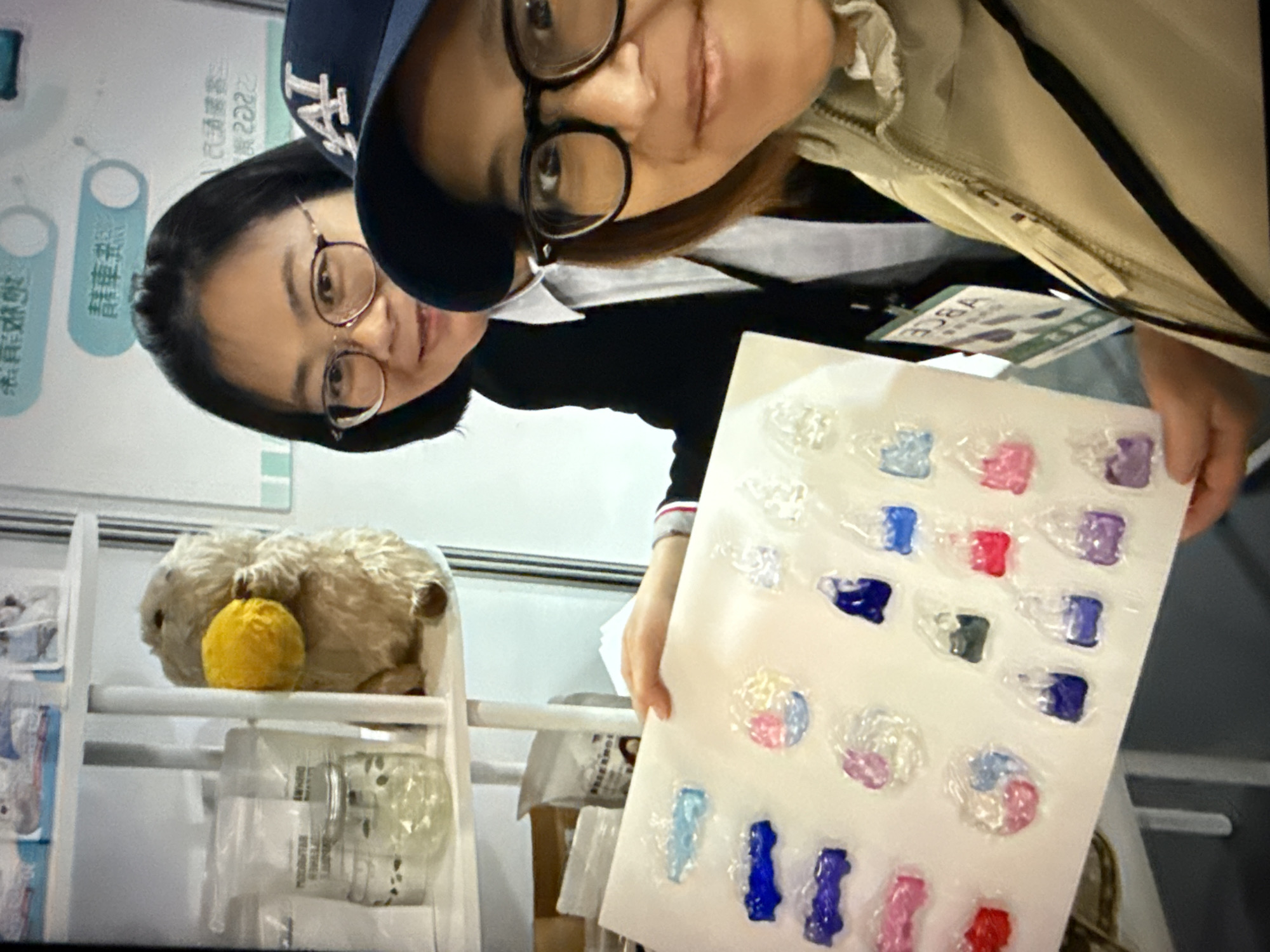 Two women are taking a selfie at what appears to be a trade show or exhibition, possibly in Taipei, Taiwan, given the presence of Chinese text in the background. The woman in the foreground, wearing glasses and a beige jacket, is on the right side of the frame, smiling at the camera. She has a dark baseball cap on. The woman behind her, also wearing glasses and a black suit jacket over a white shirt, is to the left and smiling. She is holding up a white board with various colorful, small, translucent items, possibly candies or samples, arranged in rows. The items are encased in clear plastic and vary in shape and color, including blue, purple, pink, and red. In the background, shelves are visible with some products and a stuffed toy of a marmot holding a yellow object. One sign in the background has Chinese characters and what looks like the year "2022". Another sign below it has "ABCA" printed on it vertically. The overall environment suggests a commercial or promotional setting.