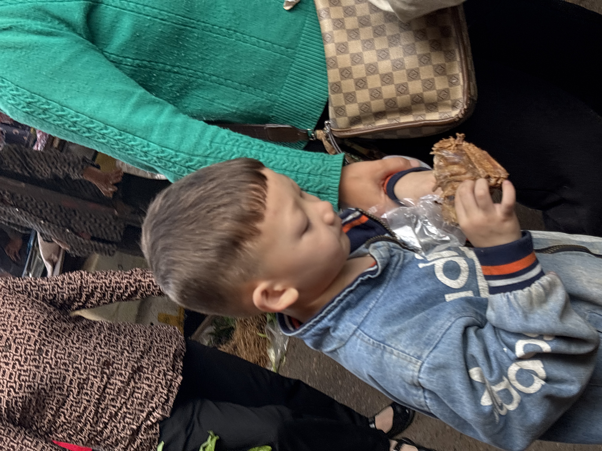 A child in a blue denim jacket with white lettering and striped cuffs is holding food wrapped in plastic. The child's left hand is supporting the food, while the right hand holds it up. The child is being assisted by someone wearing a green knit sweater and a beige, patterned shoulder bag. The person's left hand is helping to hold the food. To the left of the child is a woman wearing a brown patterned dress and black pants. Black shoes are visible on her feet.