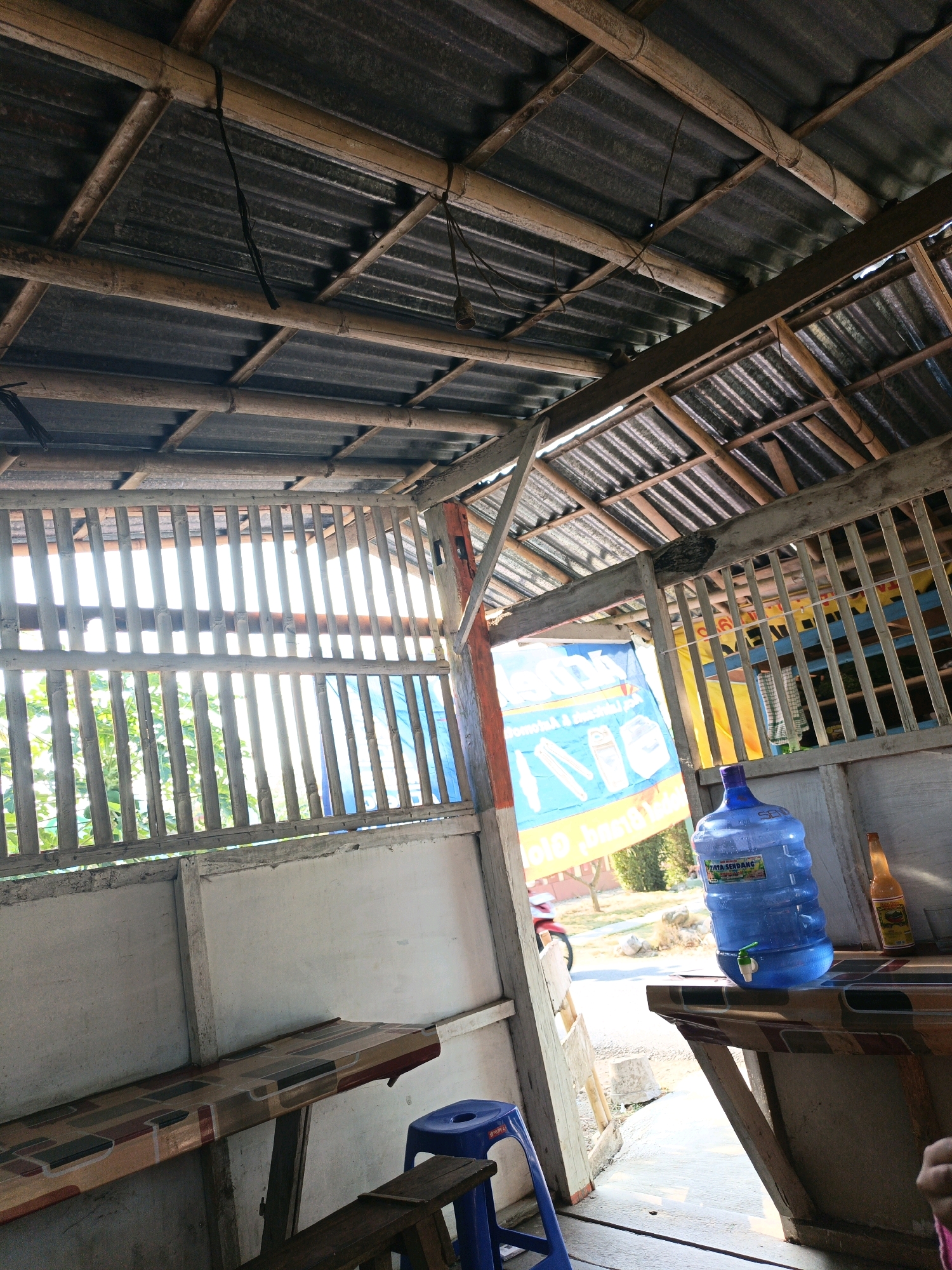 The image depicts a simple outdoor seating area under a corrugated metal roof supported by bamboo beams. The roof is angled and appears to be constructed with a combination of bamboo and metal. Vertical bamboo poles form a slatted wall on the left, allowing sunlight to filter through, illuminating a green leafy plant outside. A blue banner with white graphics and text is visible behind the bamboo poles, and a red motorcycle is partially visible in the background.
In the foreground, a long table with a colorful, patterned laminate top is supported by wooden legs. On the right side of the image, a large blue water dispenser with a green tap sits on a table. Next to the water dispenser is a bottle of amber liquid with a yellow label. A small blue plastic stool is positioned near the table. The ground appears to be concrete. The scene is consistent with an open-air food stall or roadside resting area in a rural or semi-rural setting. Given the reference to Buayan, Indonesia, this would be a typical roadside establishment in that region.